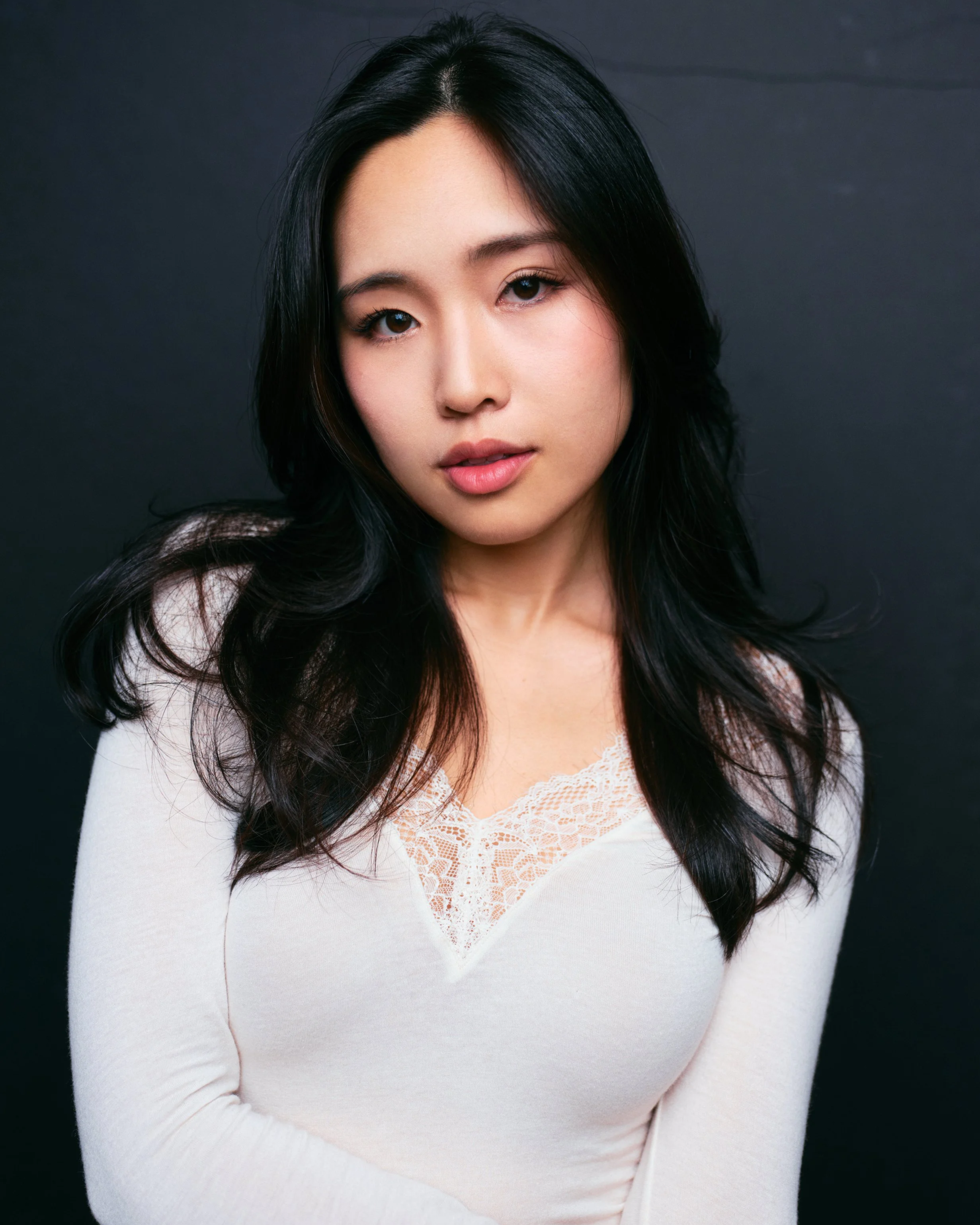 Portrait of a young woman with long black hair, wearing a white top with lace detail, against a dark background.
