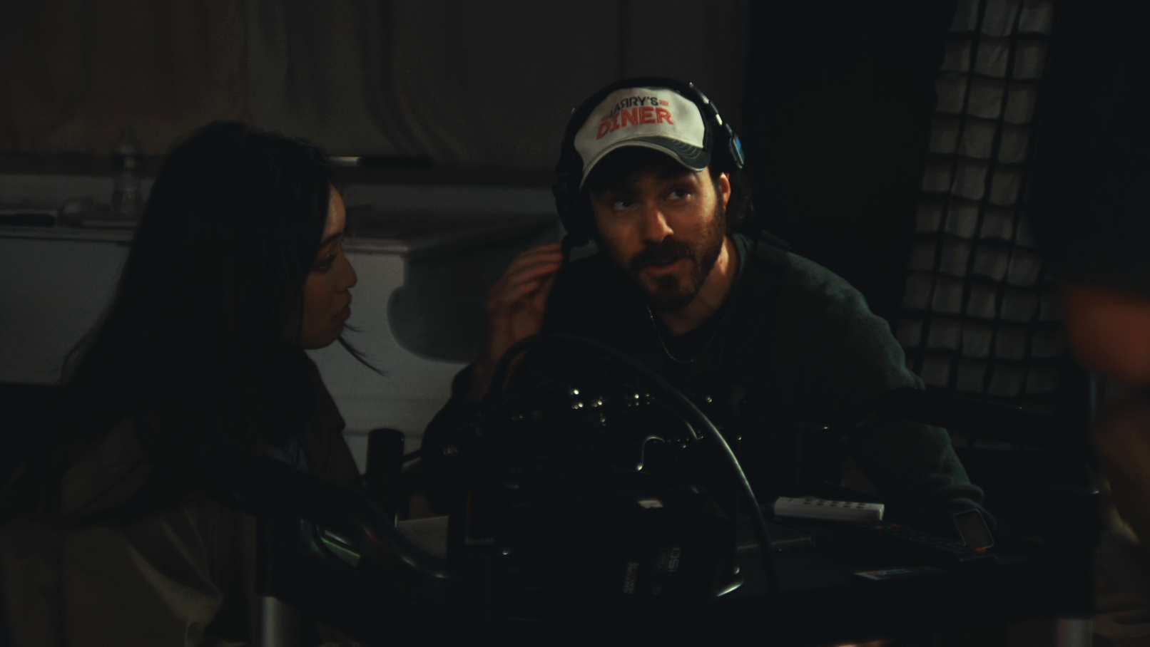 A man wearing a white cap with red text, a gray sweater, and headphones sitting at a control panel with a woman in dark clothing in a dimly lit room.