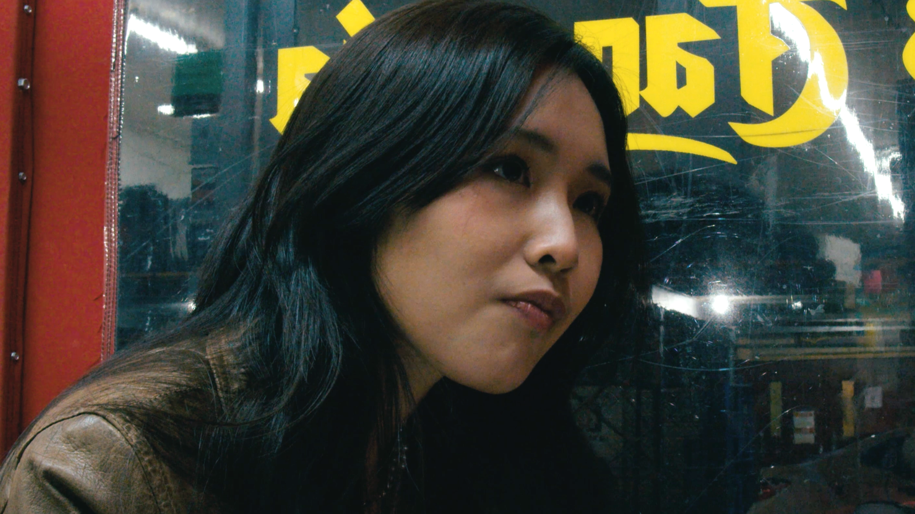 Portrait of a young woman with long black hair, wearing a tan jacket, sitting near a window with yellow lettering behind her.