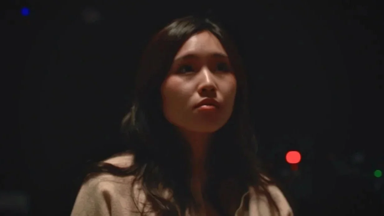 A woman with long dark hair looking towards the camera in a dark setting.