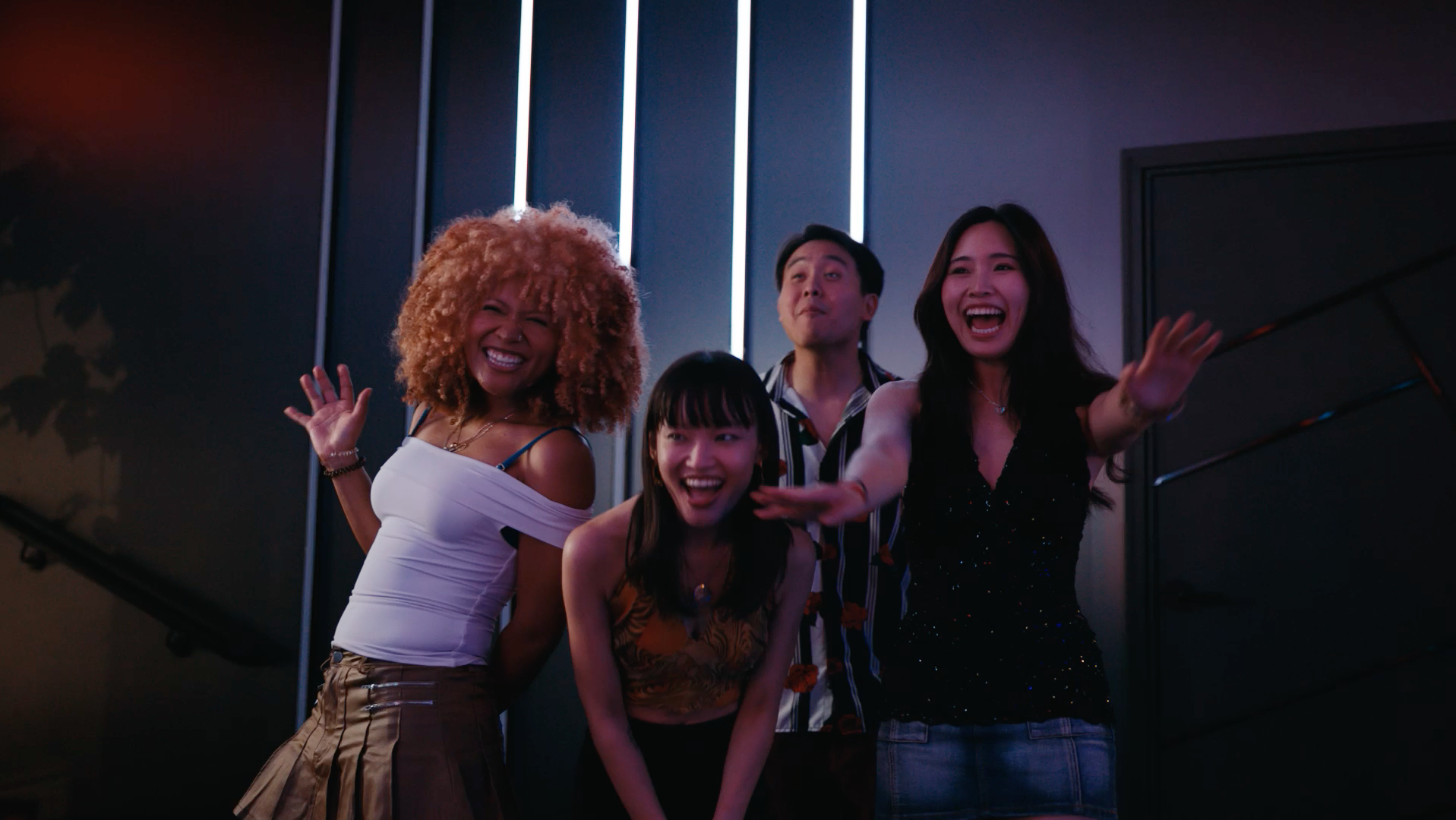 Three women taking a selfie in a colorful, neon-lit nightclub or bar. One woman has curly blonde hair, another has long dark hair and is making a pouty face, and the third woman has long black hair and is making a peace sign. A man with glasses, blurred in the foreground, is looking at them.