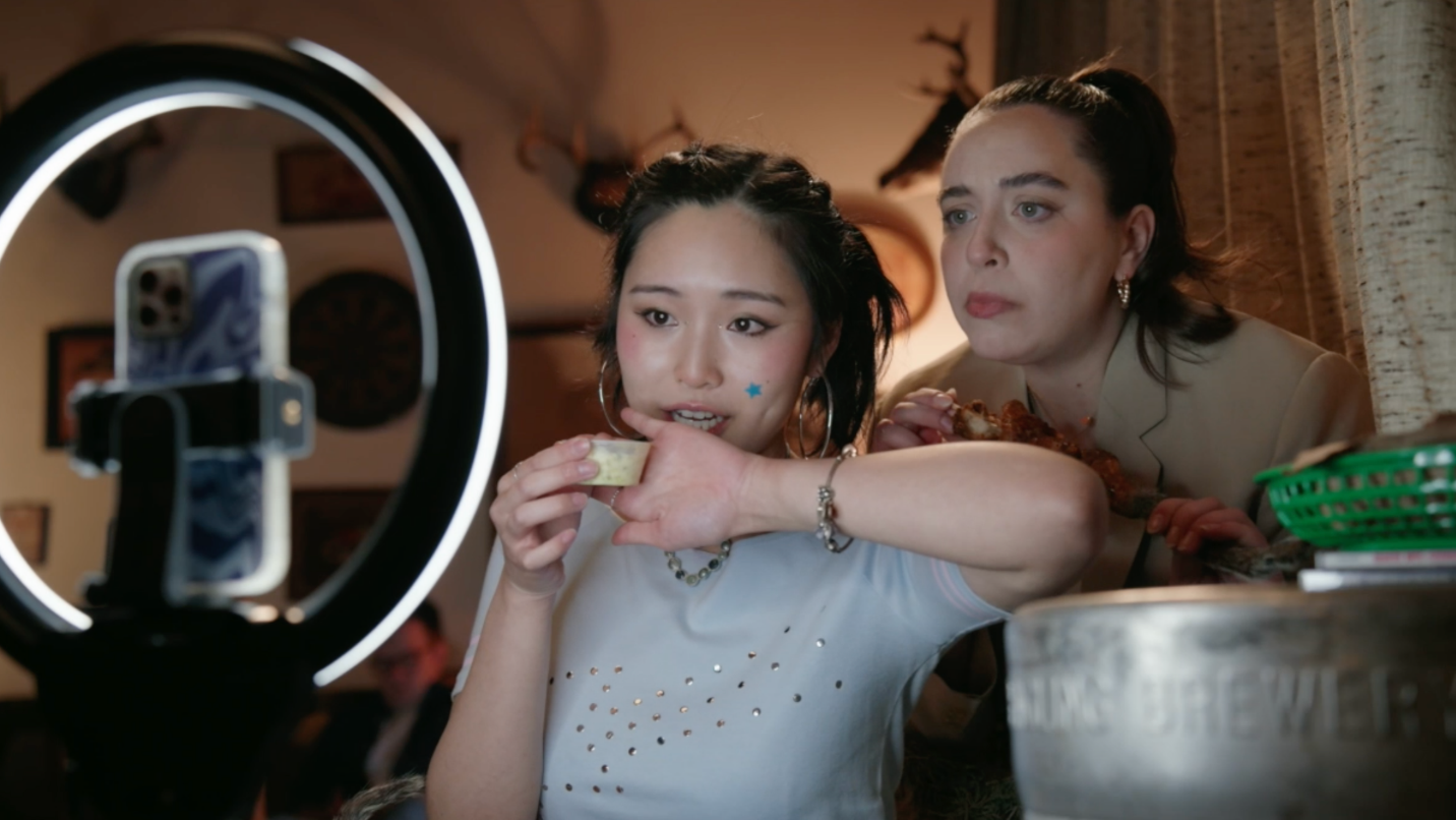 Two women look at a smartphone mounted on a ring light, with one woman eating food and the other watching, in a cozy indoor setting.