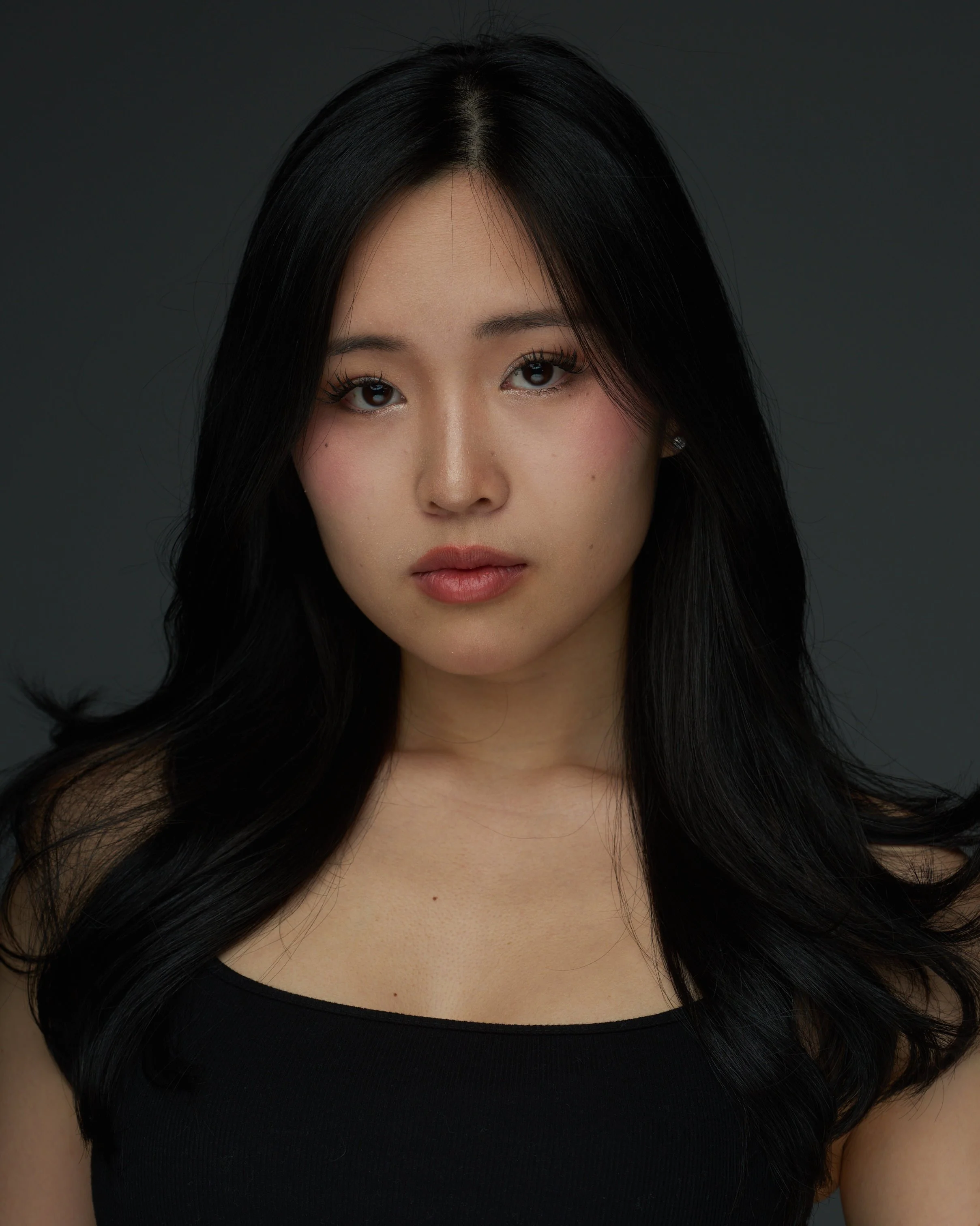 Portrait of a young woman with long black hair, wearing a black top, against a dark background.