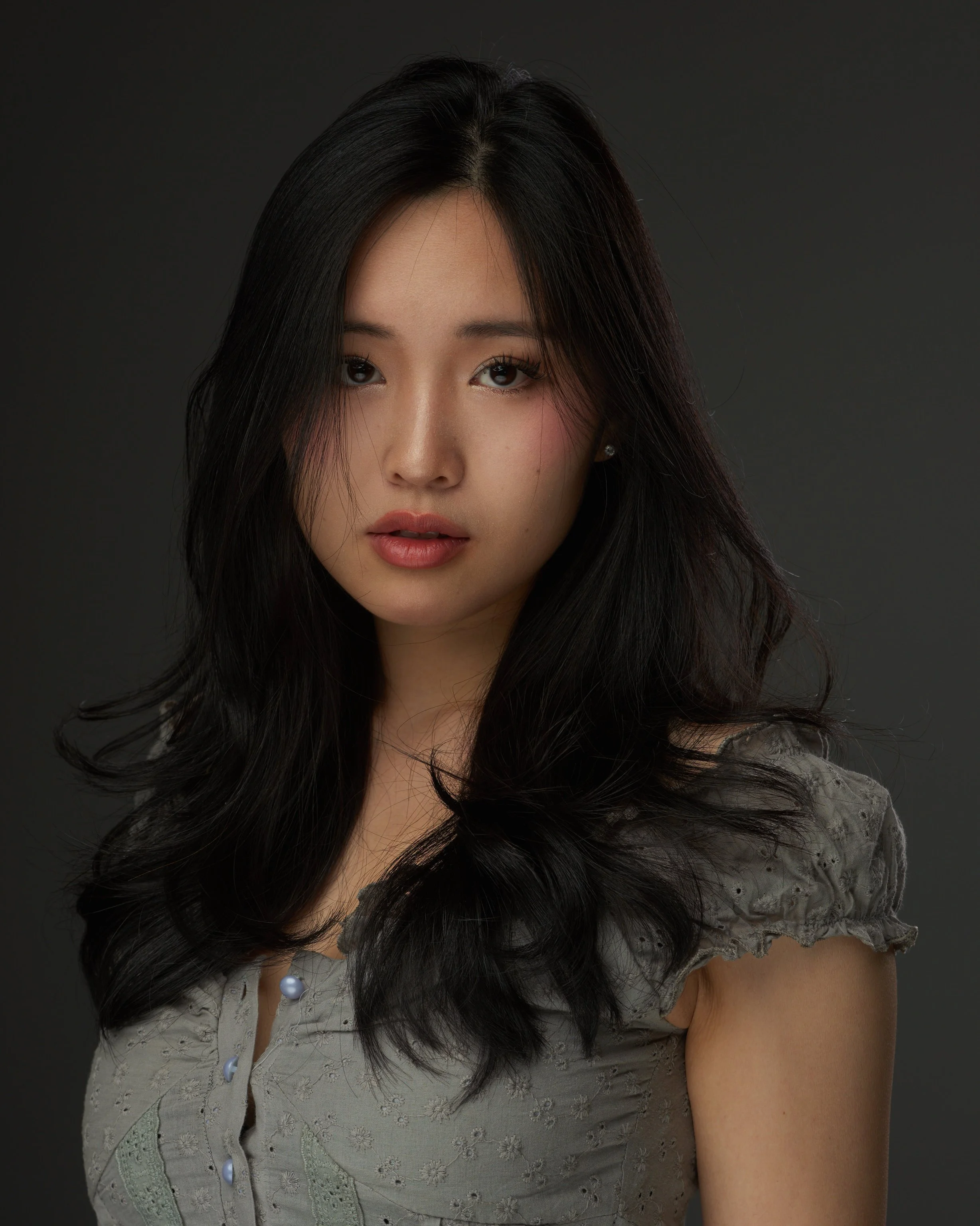 Portrait of a young woman with long, wavy black hair, looking directly at the camera, wearing a light gray dress with ruffled sleeves, against a dark background.