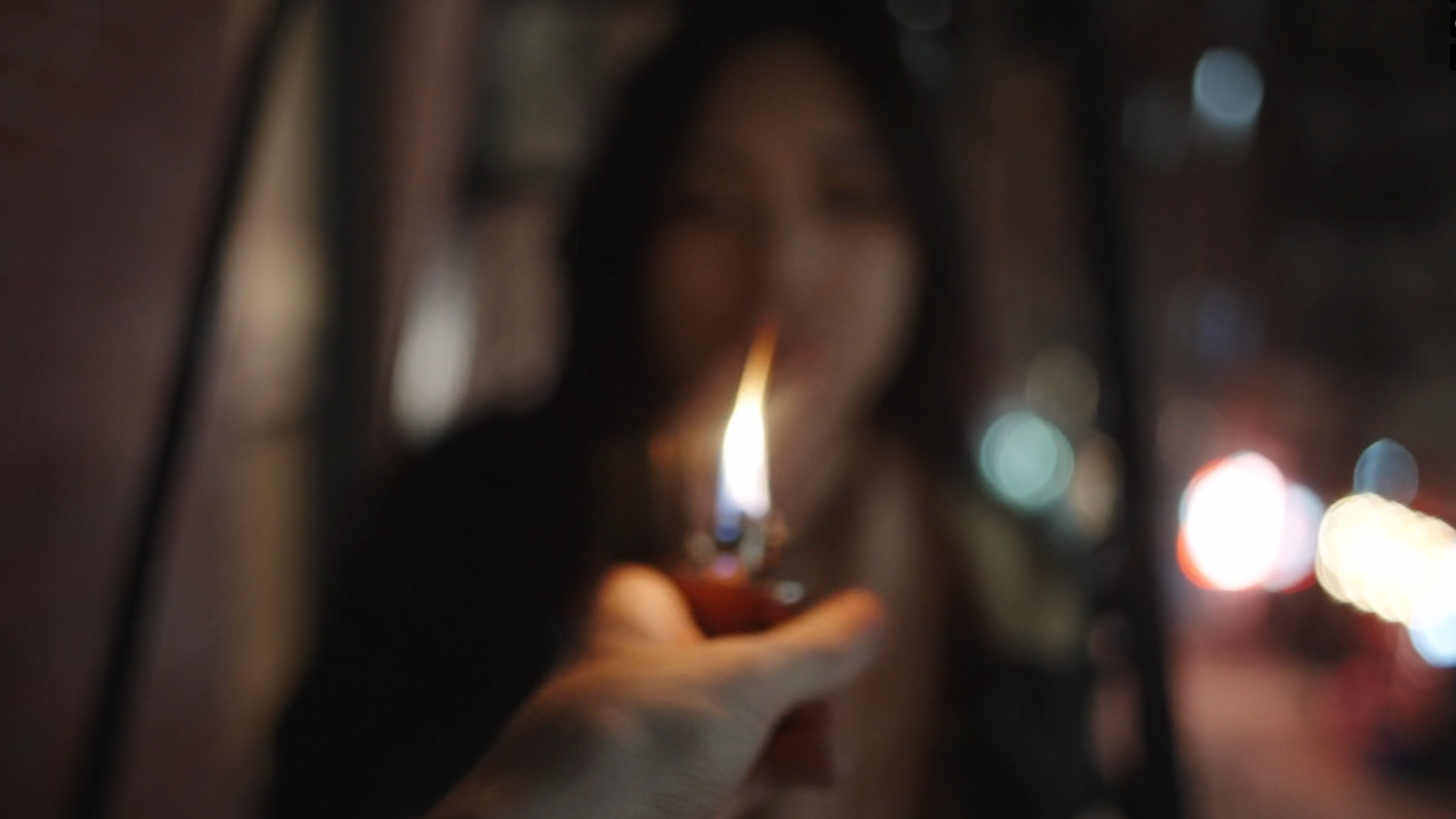 A person holding a lighter with flame near a window at night, with bokeh city lights in the background.