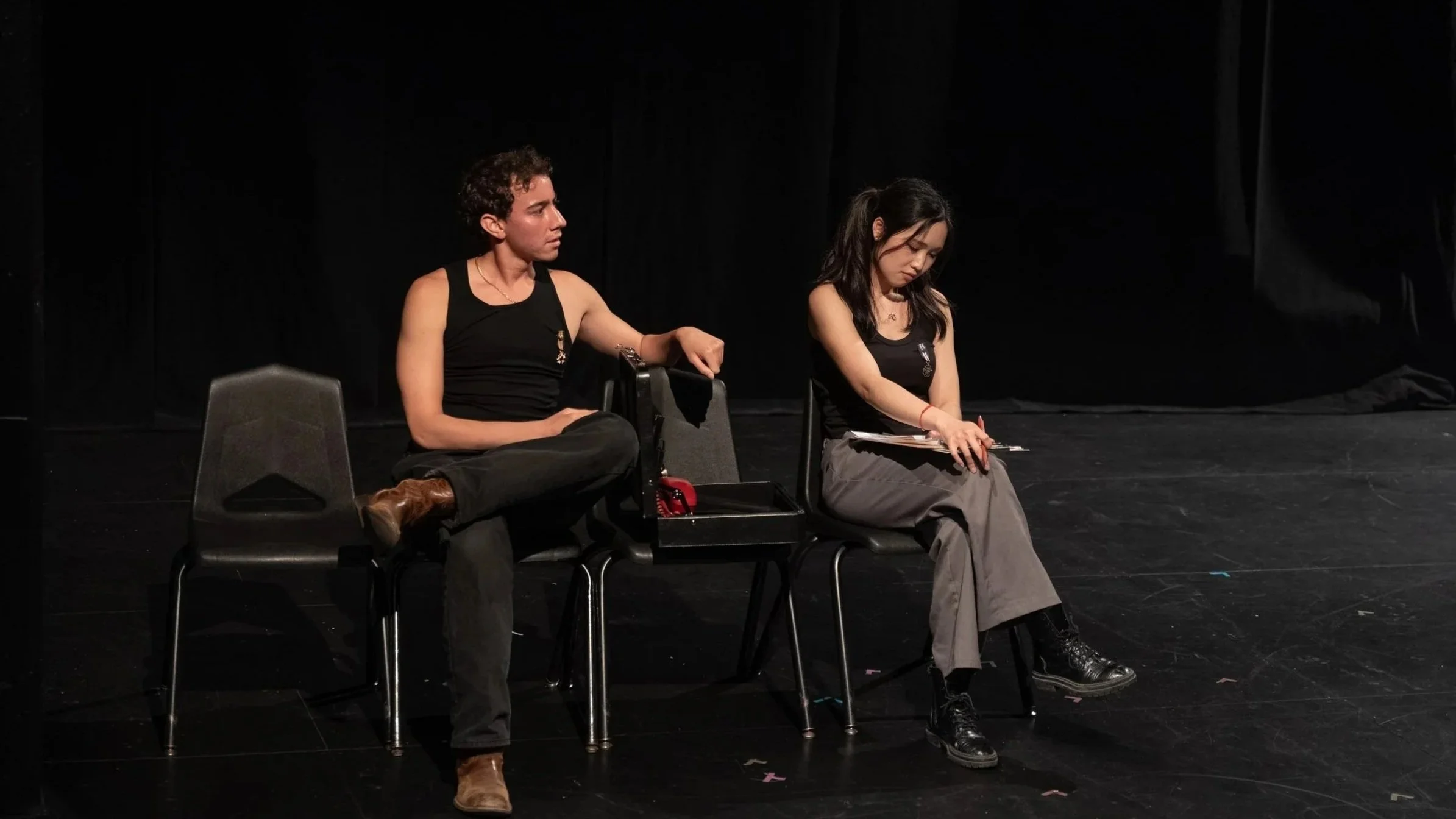 Two actors sitting on chairs on a stage during a theatrical performance, one male and one female, both wearing black tank tops and dark pants, with dark curtains in the background.