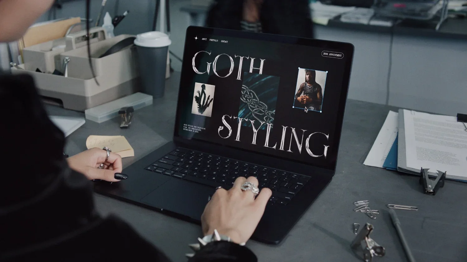 A person designing a gothic-themed page titled 'Goth Styling' on a laptop in a workspace with office supplies, papers, and a coffee cup nearby.
