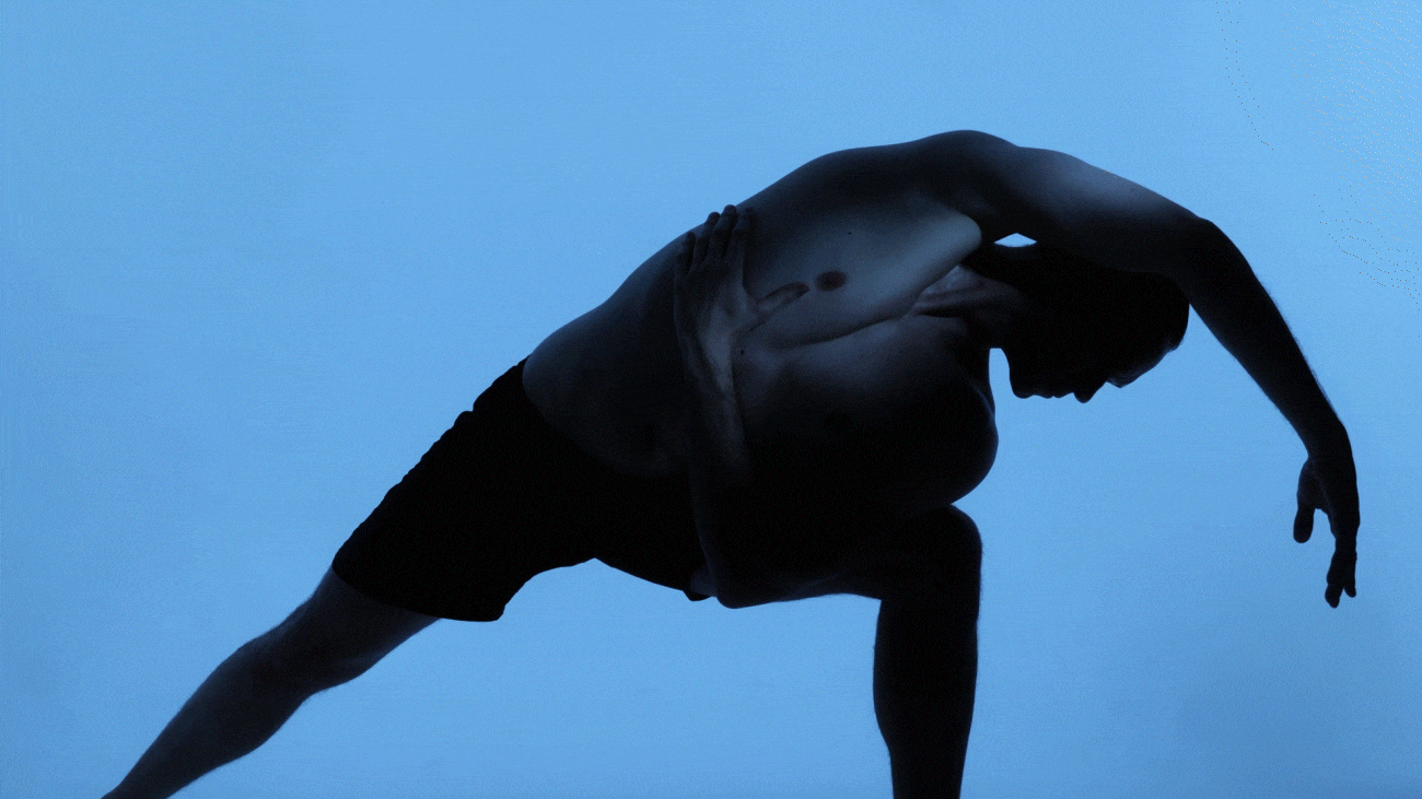 A person with a muscular build is in a yoga or dance pose against a blue background. They are shirtless, wearing black shorts, with one arm wrapped around their back and the other extended downward, head bent forward.