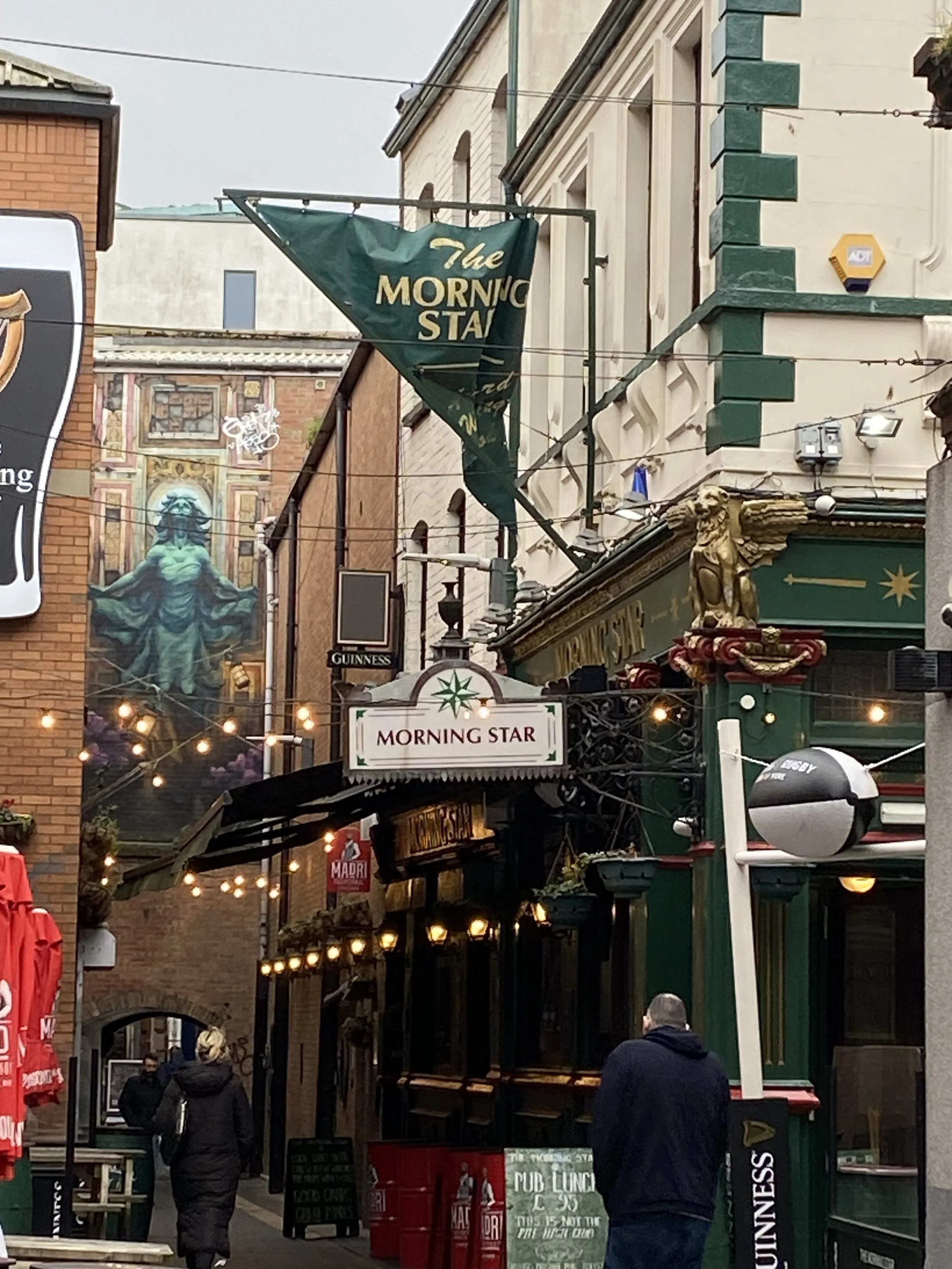 Street scene featuring the Morning Star pub, hanging signs, string lights, and people walking in a city alley.