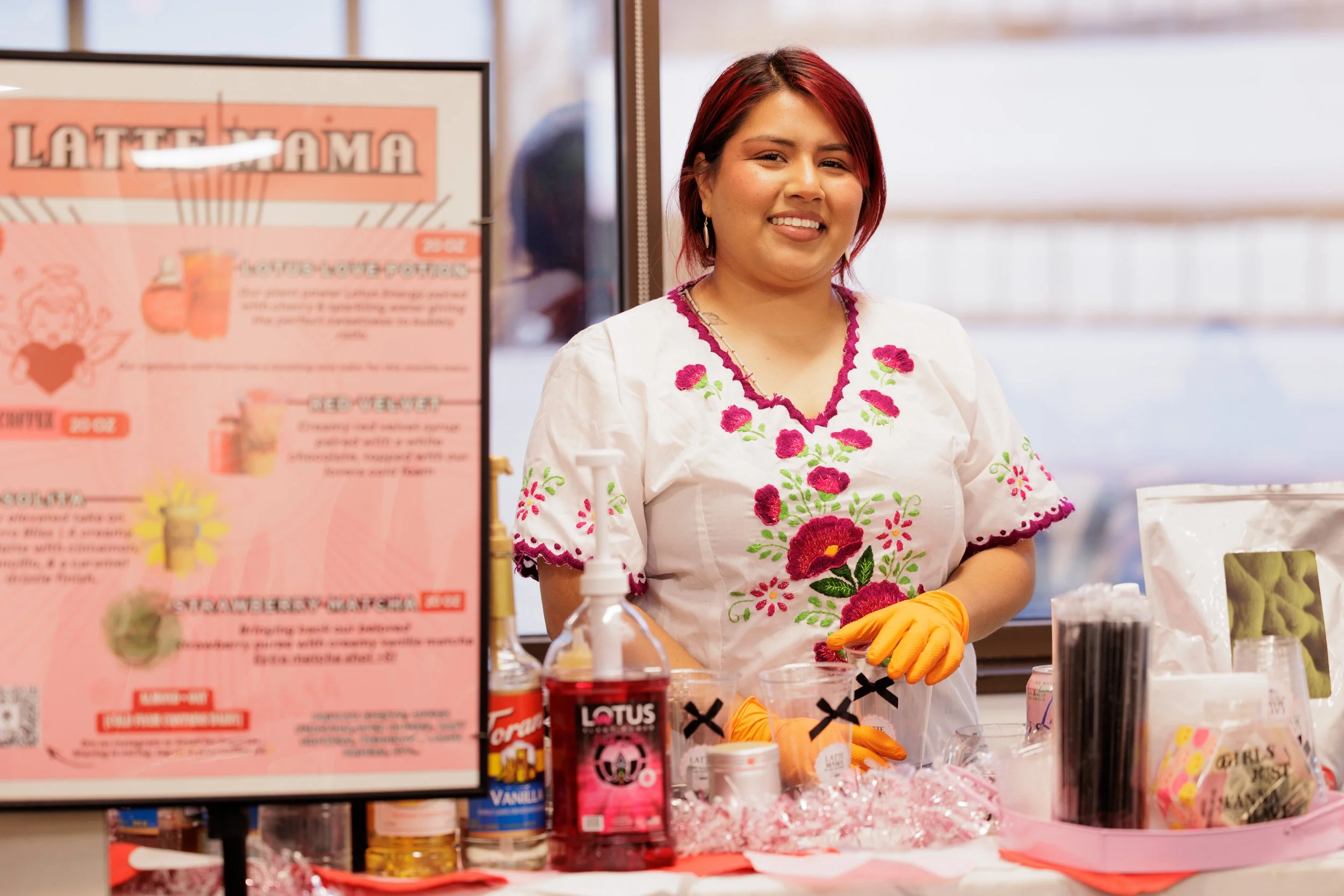 A woman makes an order at a cafe counter, with two women standing in front of her, one in a black shirt and the other in a yellow top, inside a modern cafe with a refrigerator and food items on the counter.