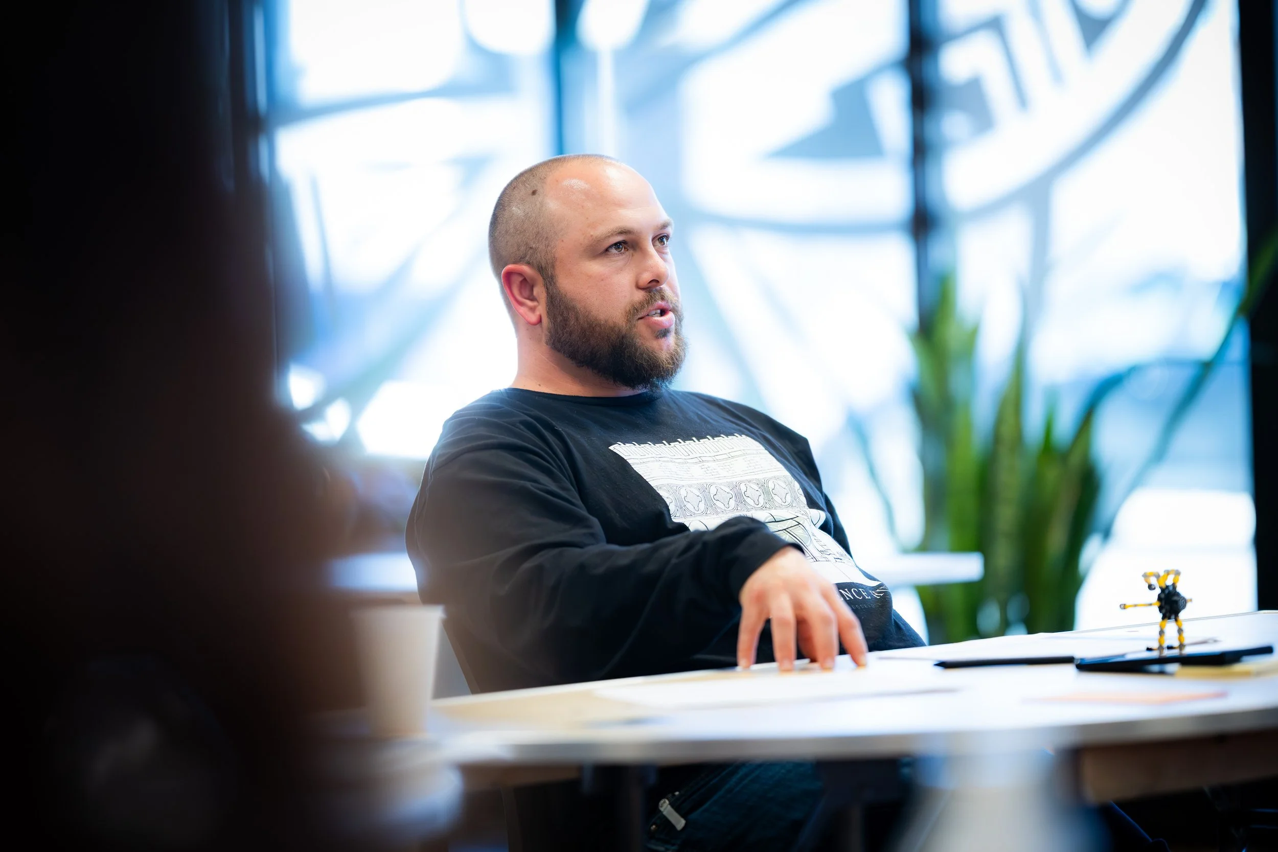 Two men are sitting at a table, looking at a laptop. One man is gesturing and the other is wearing a hat and holding a small iron on a wooden base.