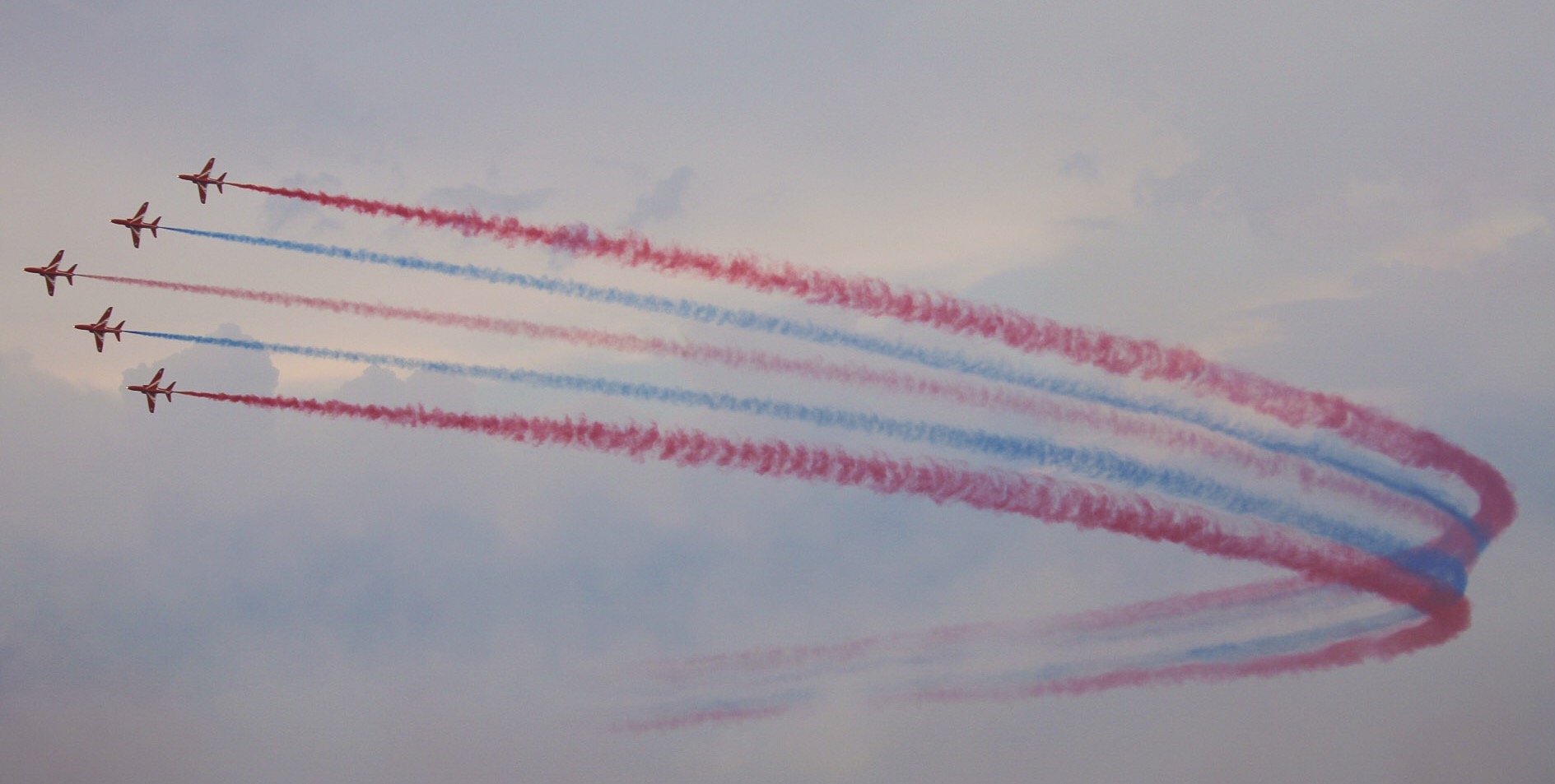 Five of the famous Red Arrows performing aerobatics with red and blue smoke trails on a cloudy sky background.