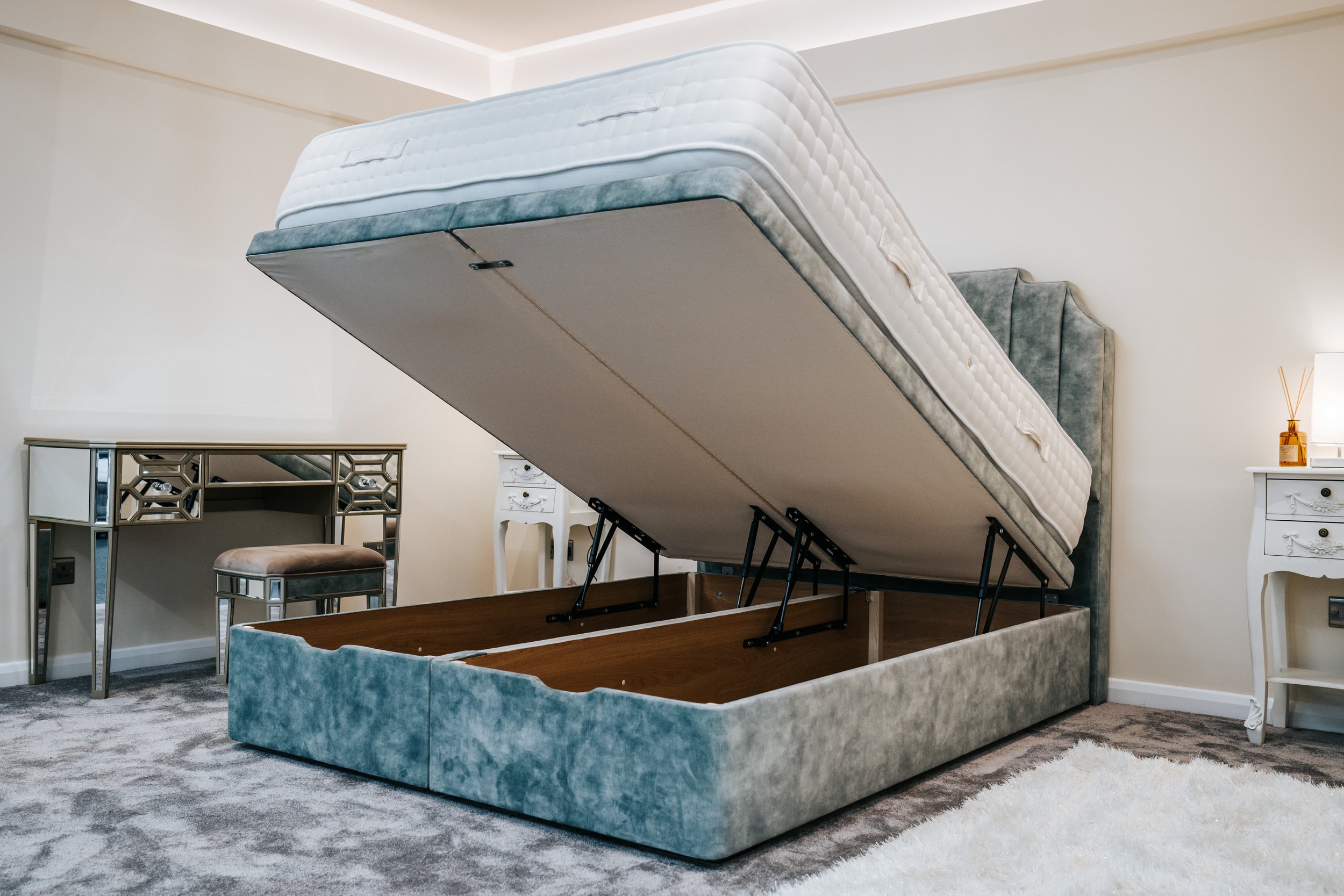 Upholstered bed with lifted mattress revealing storage space underneath, situated in a modern bedroom with mirrored vanity and soft carpet.
