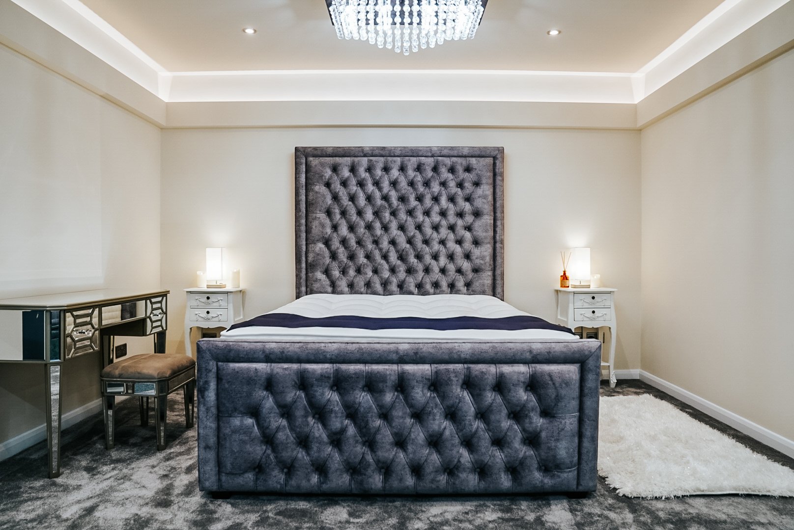 Elegant bedroom with tufted headboard, plush bed, mirrored furniture, and ambient lighting.