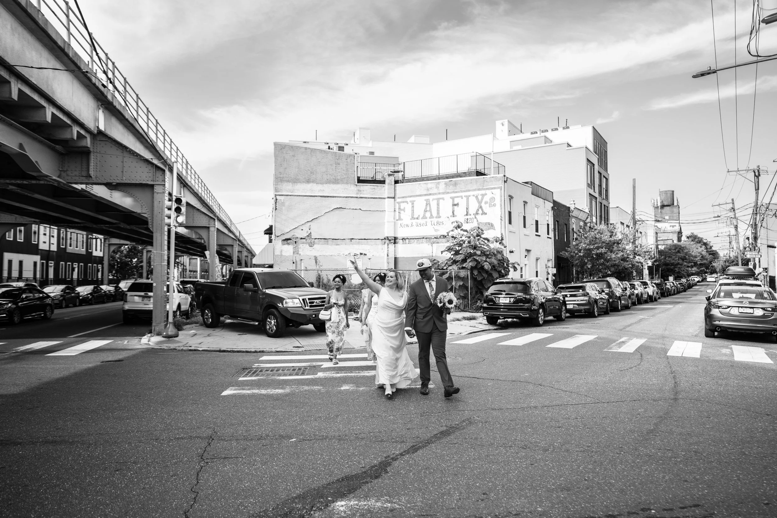 A black-and-white photo of a city street with parked cars and people crossing the road. Two women, one in a white dress holding a bouquet, walk with a man in a suit, possibly a wedding party. Buildings and an elevated train track are visible in the background, along with overhead power lines and cloudy sky.