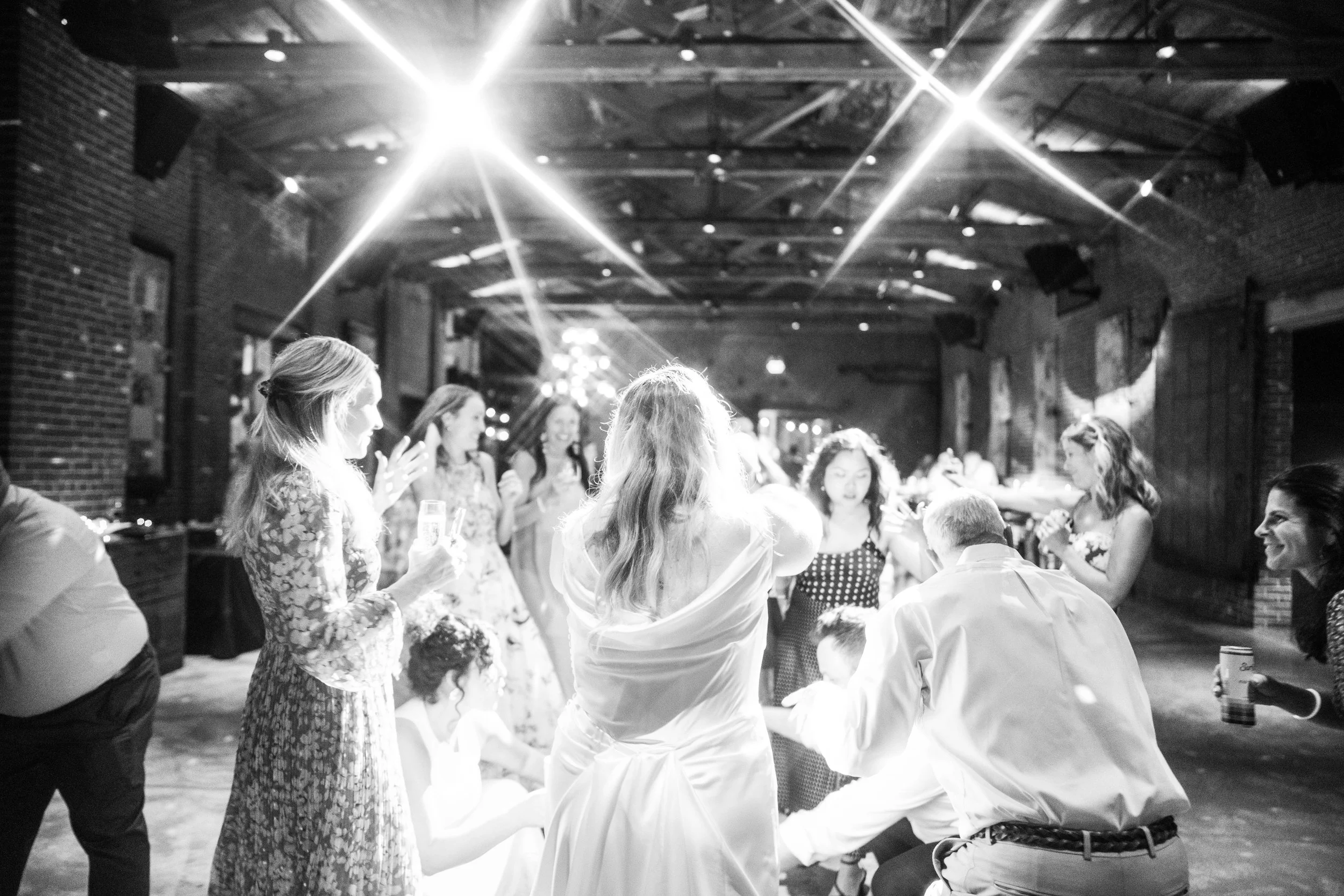 Black and white photo of a group of people, mostly women, dancing together at a social gathering or party in a rustic indoor venue with exposed brick walls and ceiling beams, bright overhead lights.