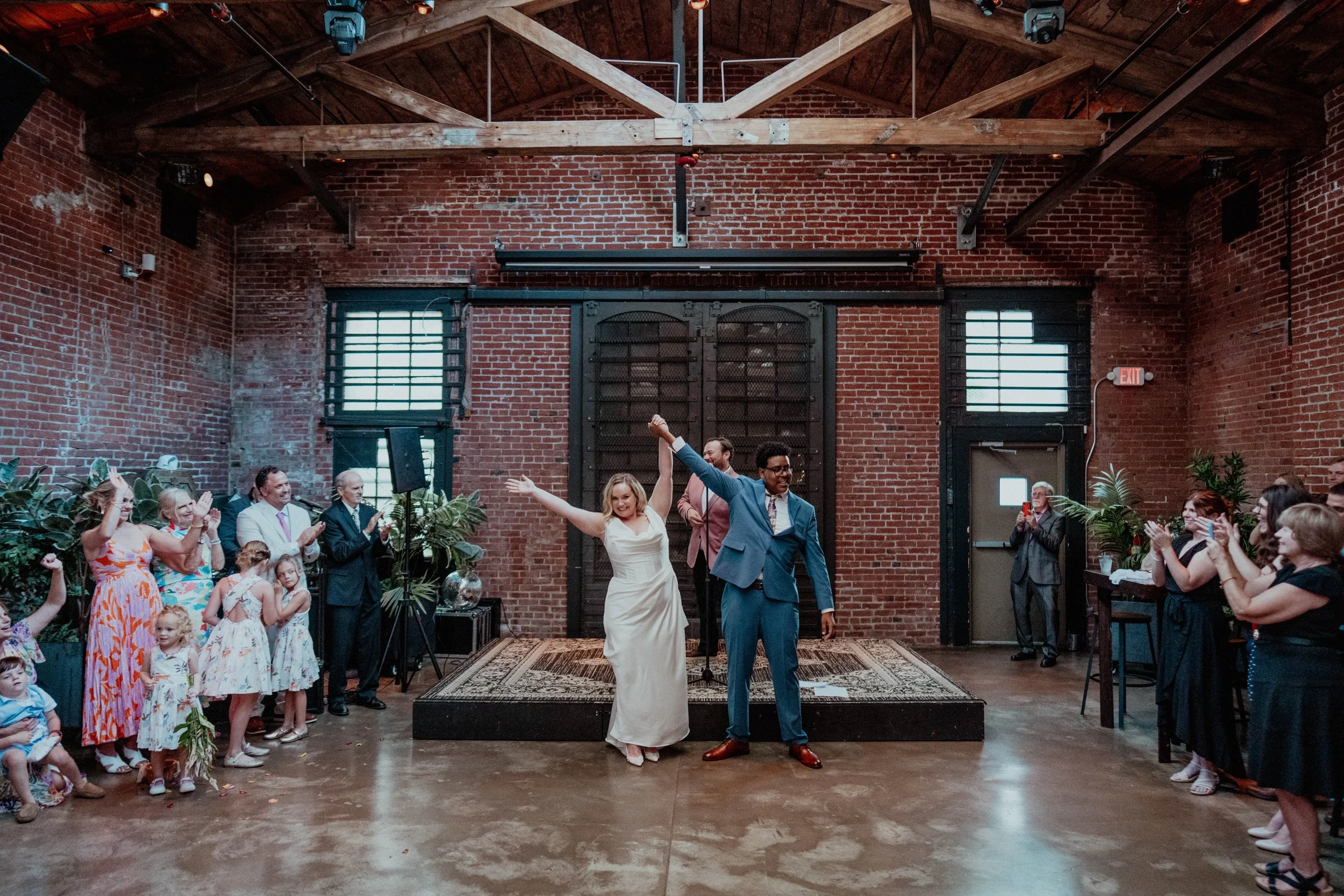 A wedding reception in an industrial-style venue with exposed brick walls and wooden beams. The bride and groom are dancing on a small stage, surrounded by guests clapping and celebrating.