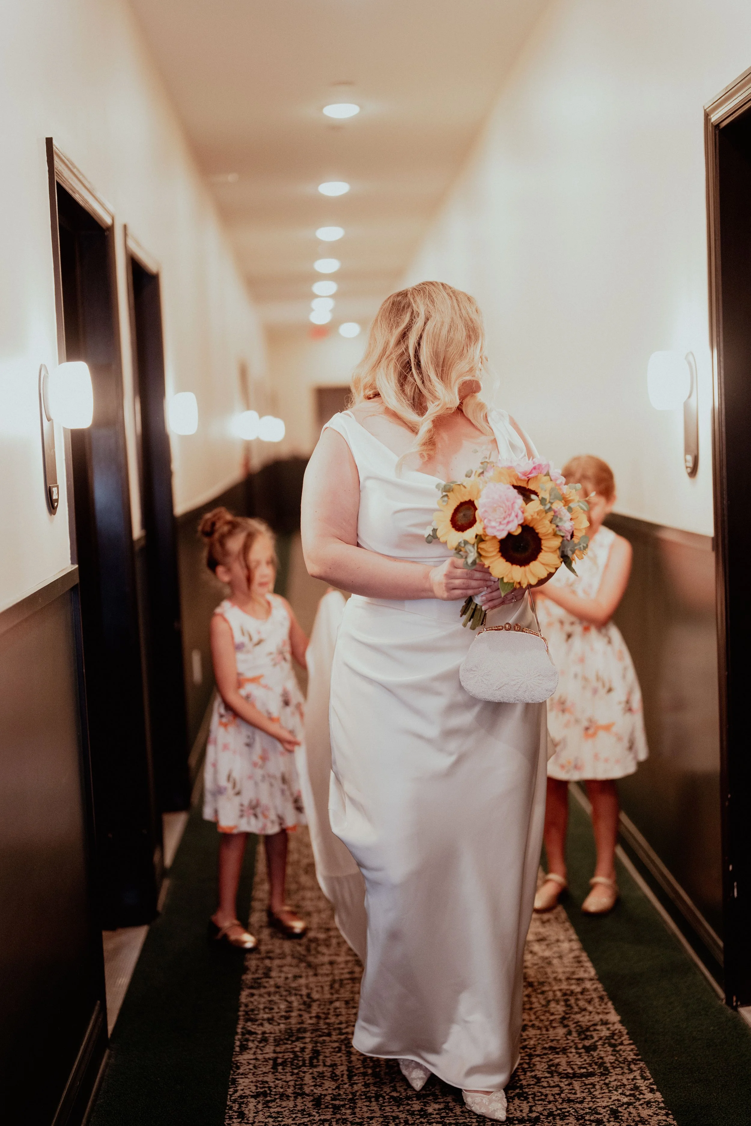 A woman in a white dress holding a bouquet of sunflowers and pink flowers, walking down a hallway with children in floral dresses following her, possibly at a wedding or formal event.
