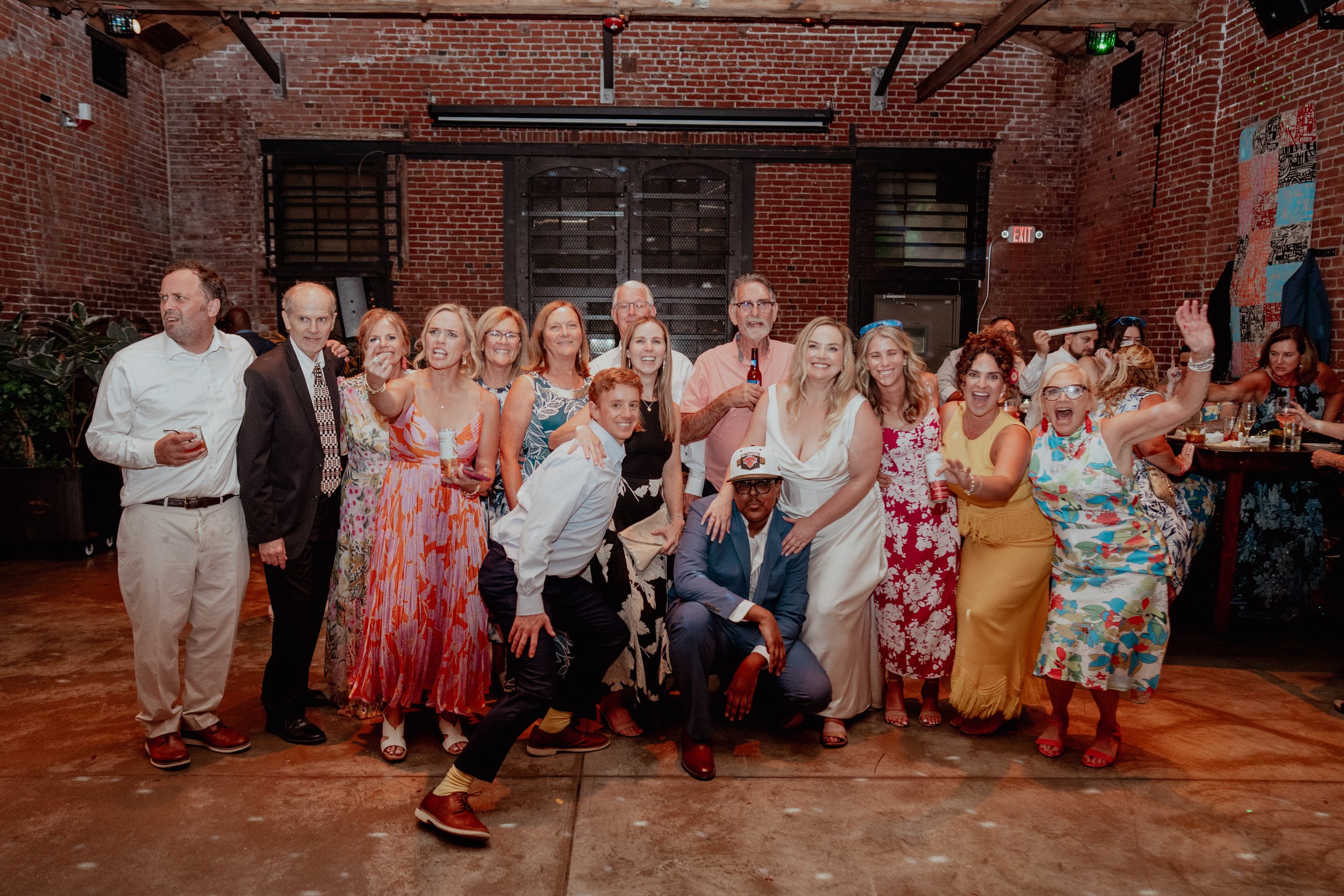 A group of diverse adults, dressed in colorful summer attire, is gathered indoors in front of a brick wall, posing for a festive photo with some smiling, making gestures, and holding drinks at a party or celebration.