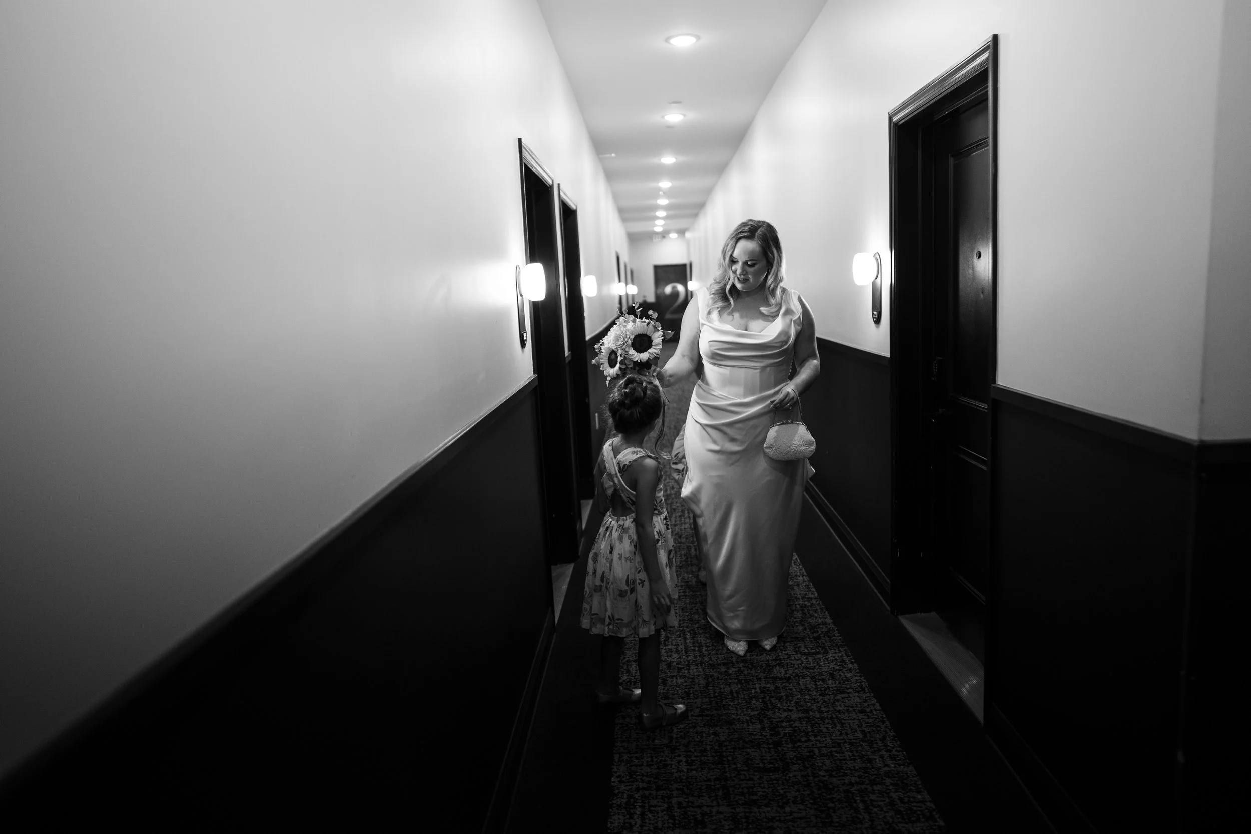 A woman in a white dress holding a bouquet of flowers and a young girl in a patterned dress walking down a hotel hallway.
