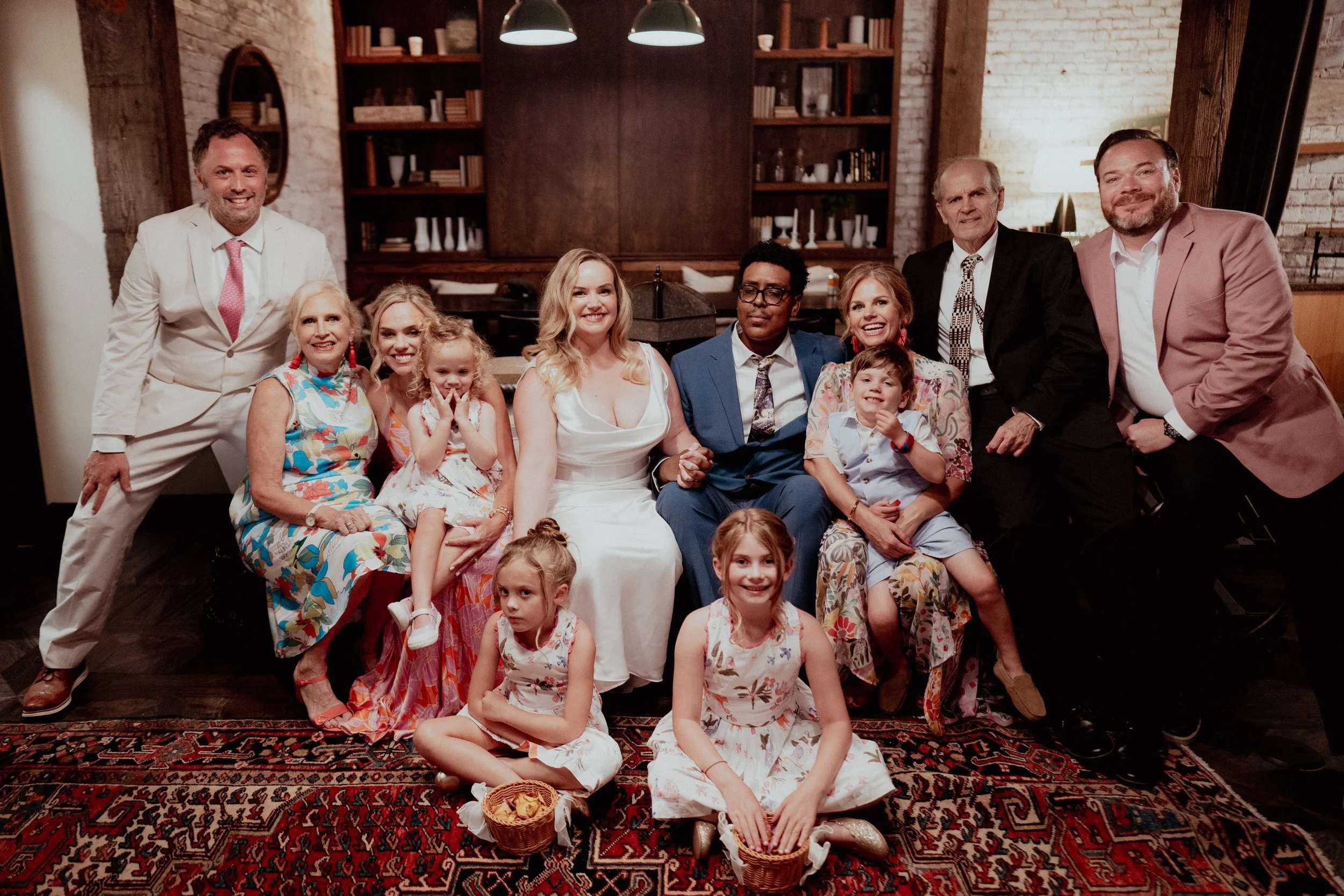 A large group of people, including adults and children, gathered in a cozy, rustic living room with brick walls and wooden furniture, posing for a group photo.