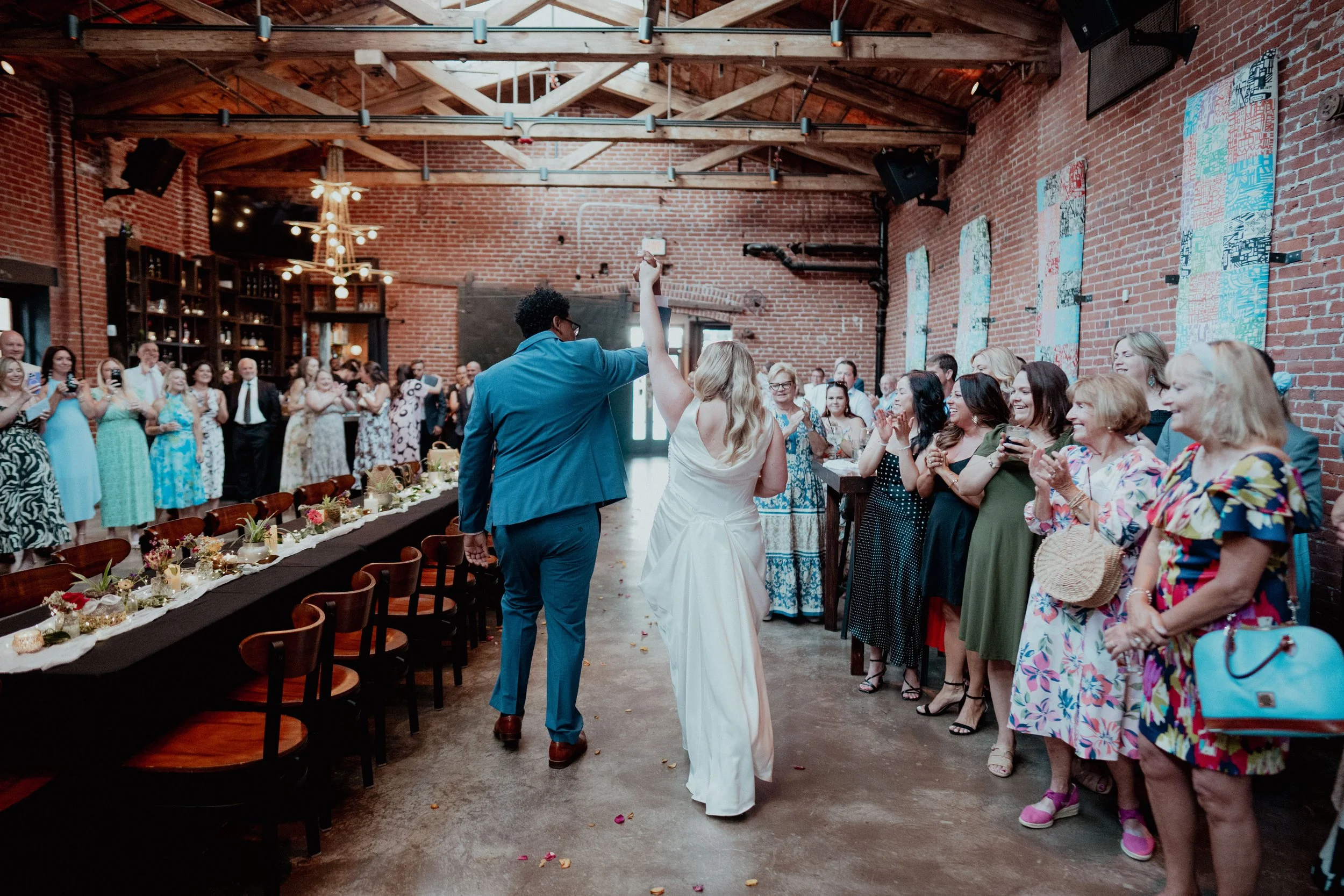 A wedding reception in a rustic-style venue with exposed brick walls and wooden beams. The bride and groom are dancing in front of an audience of guests, who are smiling and clapping. The guests are dressed in colorful dresses and suits, and the deco