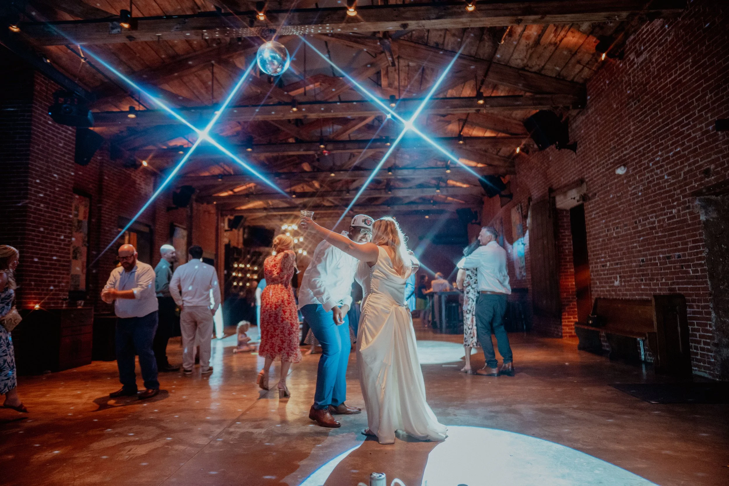 People dancing on a wooden floor in a rustic venue with exposed brick walls and a wooden ceiling; a disco ball and bright stage lights are overhead.