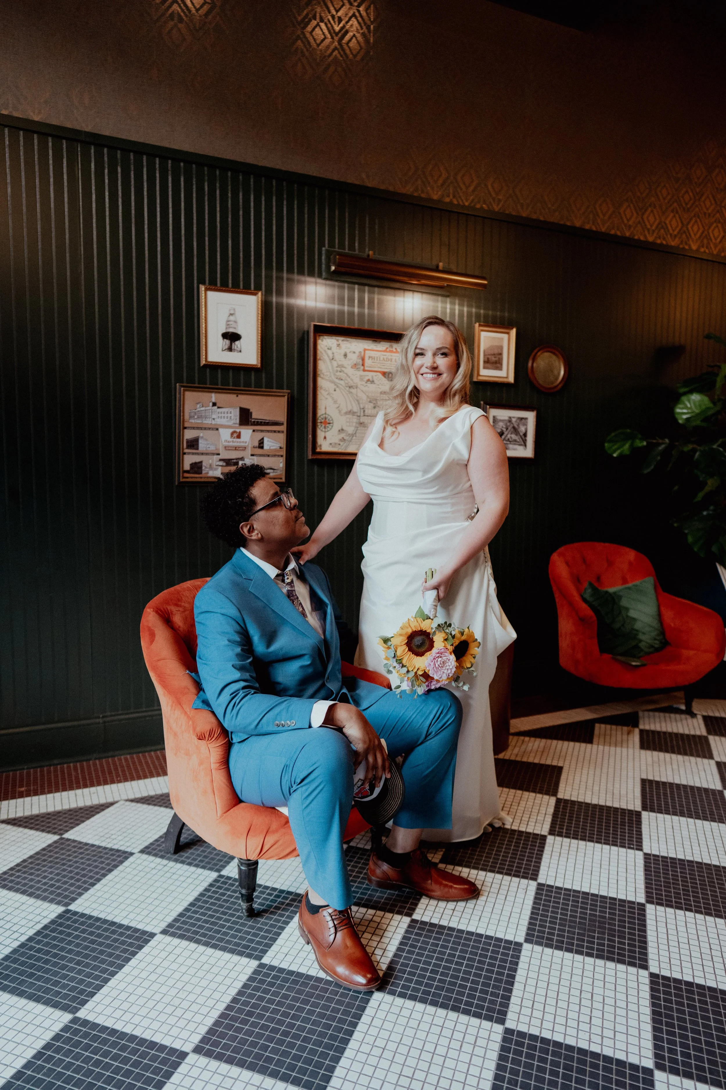 A woman in a white dress standing next to a seated man in a blue suit. The woman holds a bouquet of sunflowers and is smiling at the camera, while the man looks up at her. They are in a room with dark green paneling, framed pictures on the wall, and 