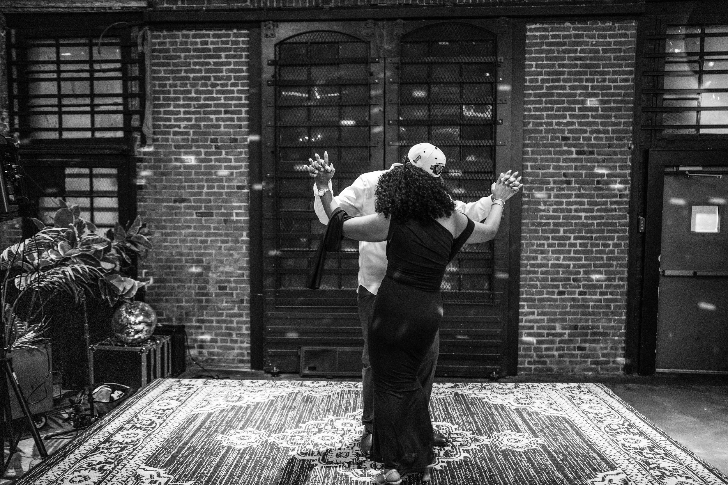A couple dancing together in a room with a brick wall background, a patterned rug on the floor, and various decorations and plants nearby.