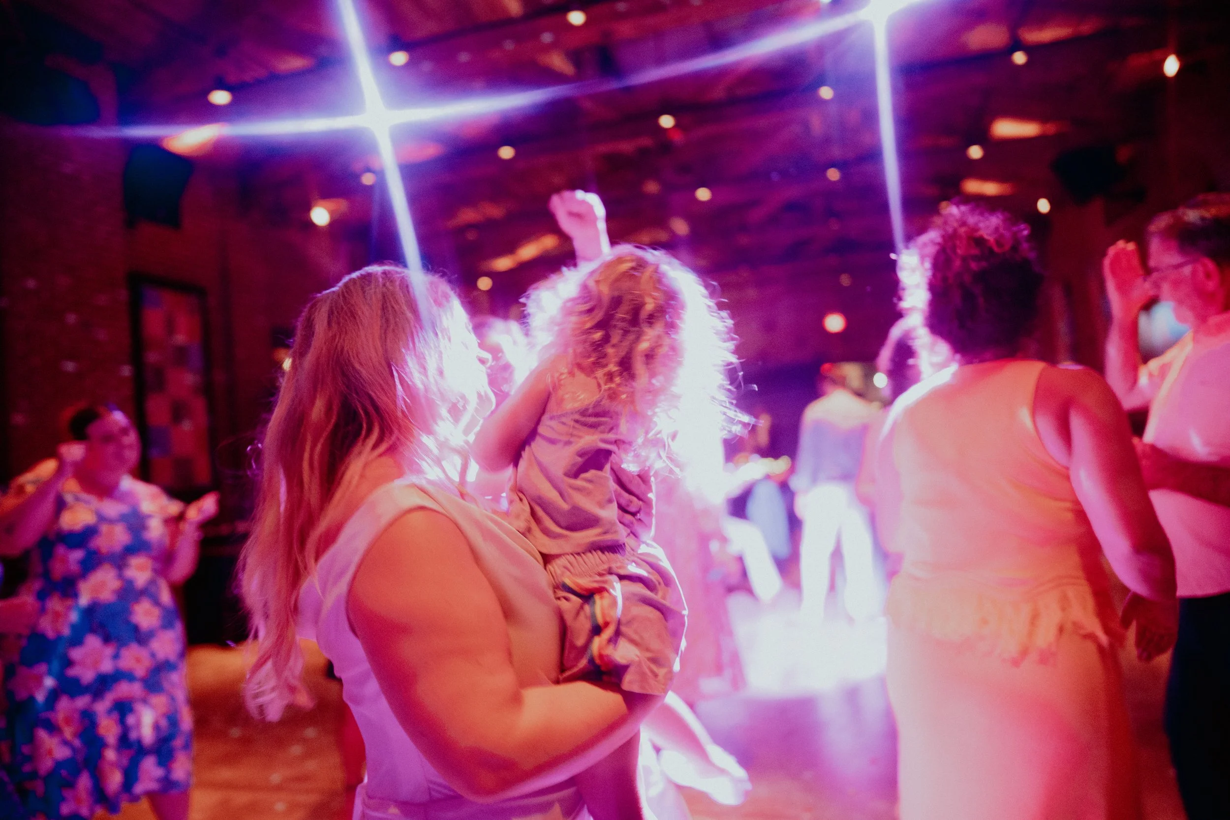 People dancing at a party or nightclub with colorful lighting and a brick wall background.
