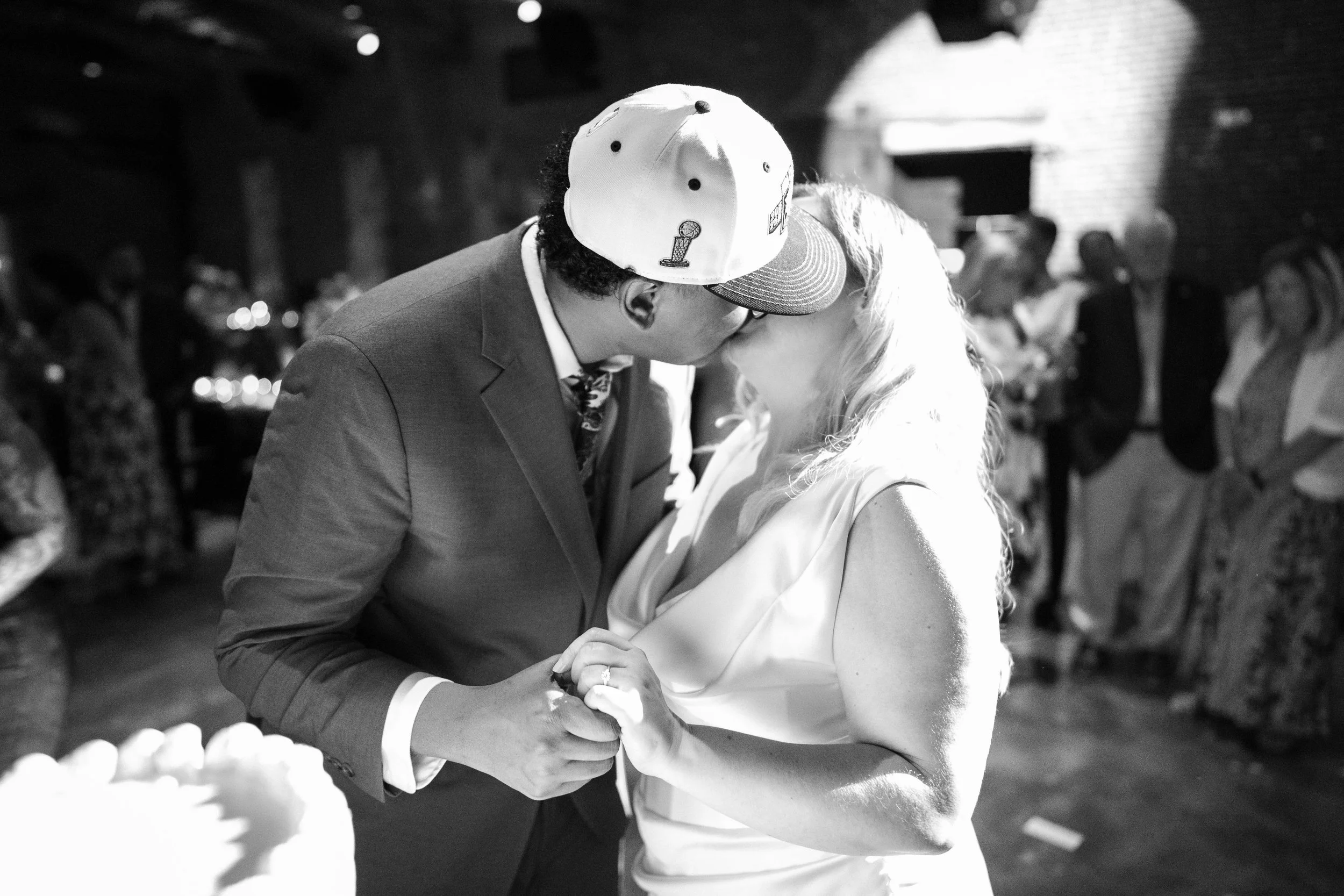 A man and woman sharing a kiss at a wedding reception, with the man wearing a suit and a baseball cap and the woman in a wedding dress.