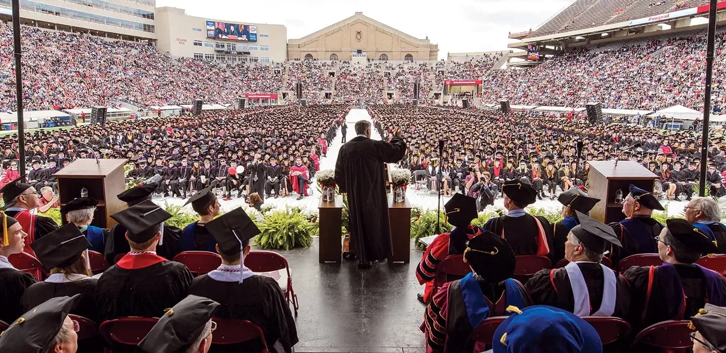 College graduation ceremony at an outdoor stadium with graduates in caps and gowns, faculty in academic regalia, and a large audience of spectators.
