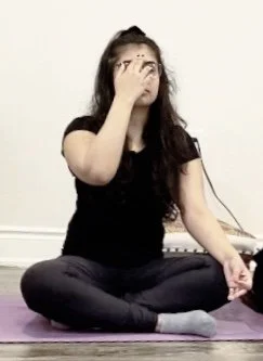 A young woman with long dark hair wearing a white top standing outdoors on a wooden balcony during sunset, with leafless trees and a tall building in the background.