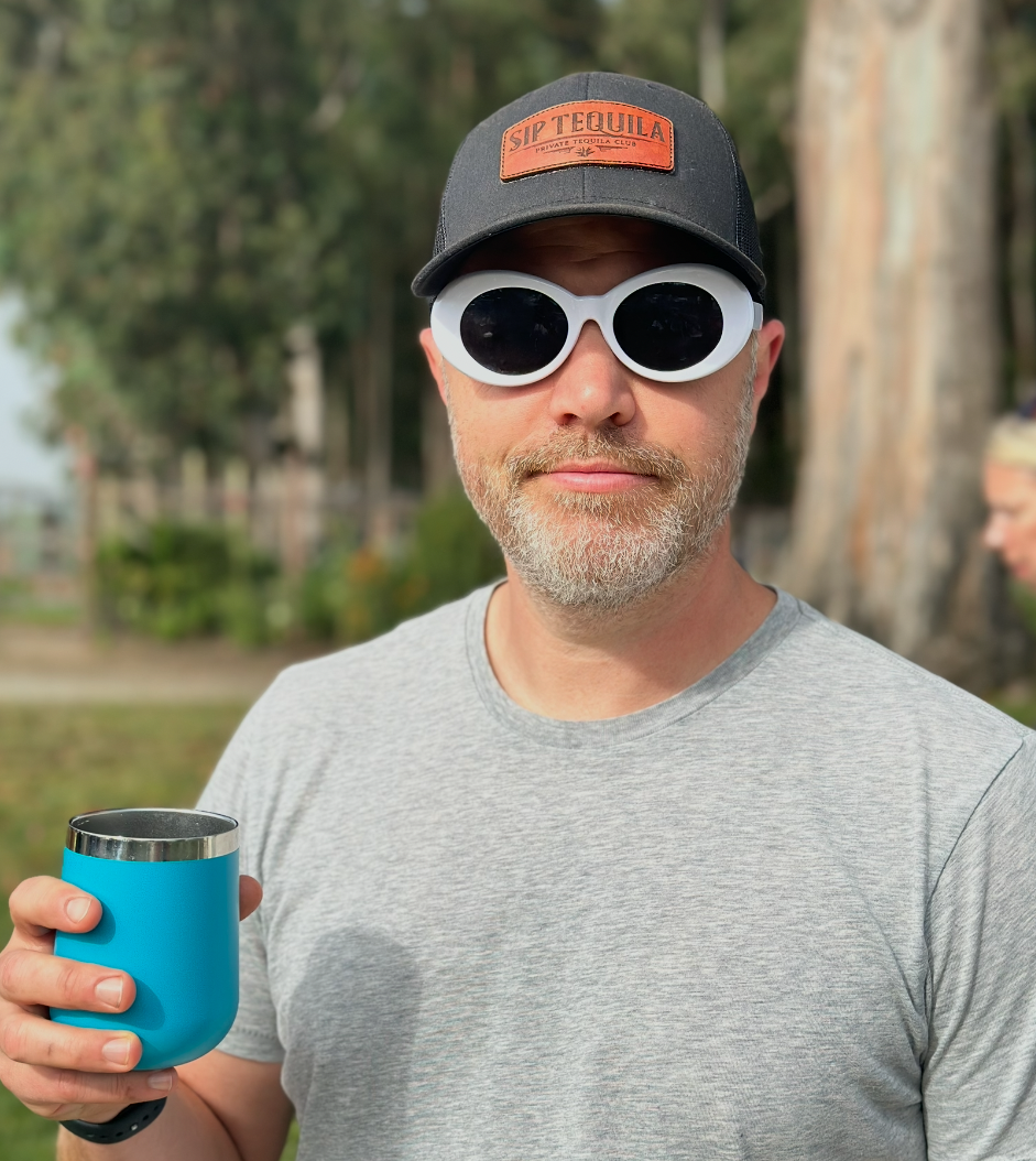 Tim Stier, Creative Director wearing a black cap labeled 'Sip Tequila', large white sunglasses, and a light gray t-shirt, holding a blue insulated cup outdoors in a park with trees in the background.