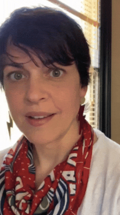 A woman with short dark hair, green eyes, wearing a white top and red patterned scarf, standing near a window with a cityscape in the background.