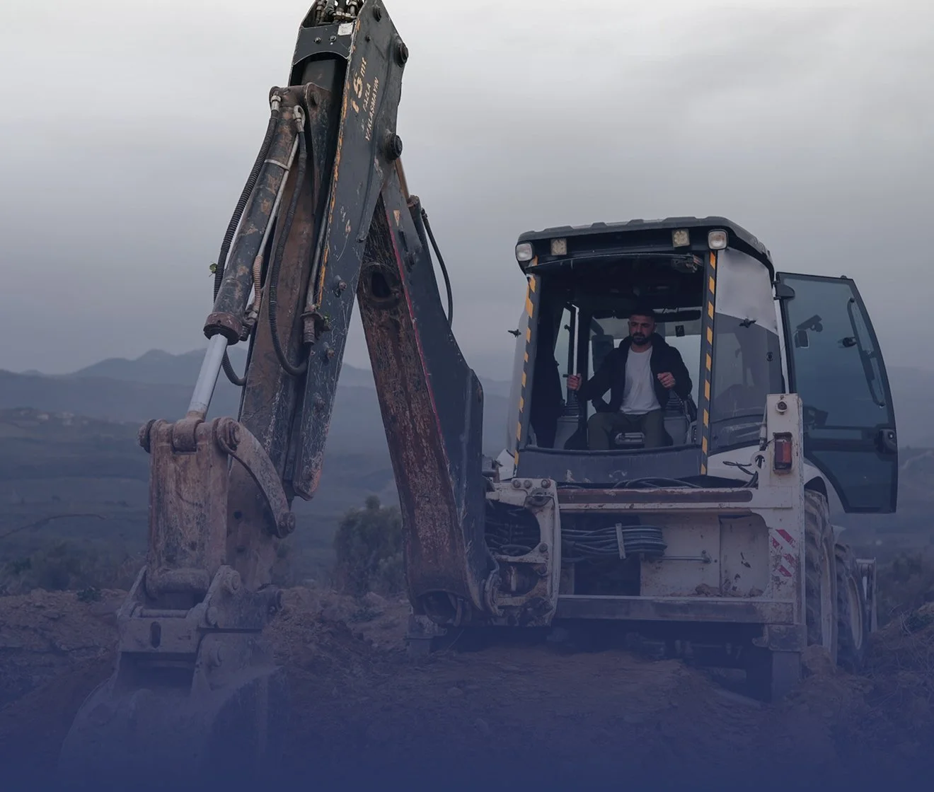 Man operating a small excavator on a construction site with mountains in the background during cloudy weather.
