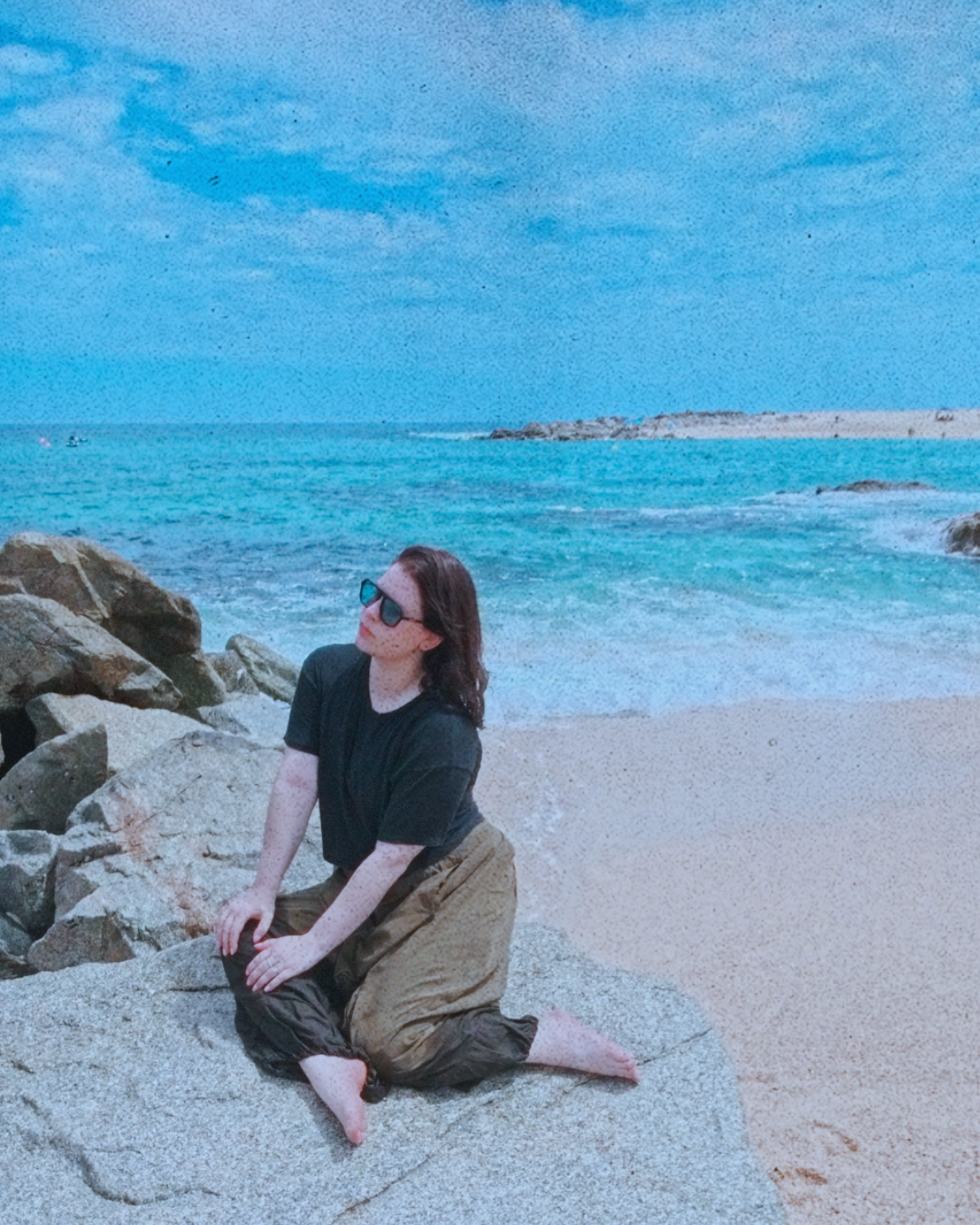 A person wearing sunglasses and a black t-shirt kneeling on a large rock by the beach, with the ocean and a cloudy sky in the background.