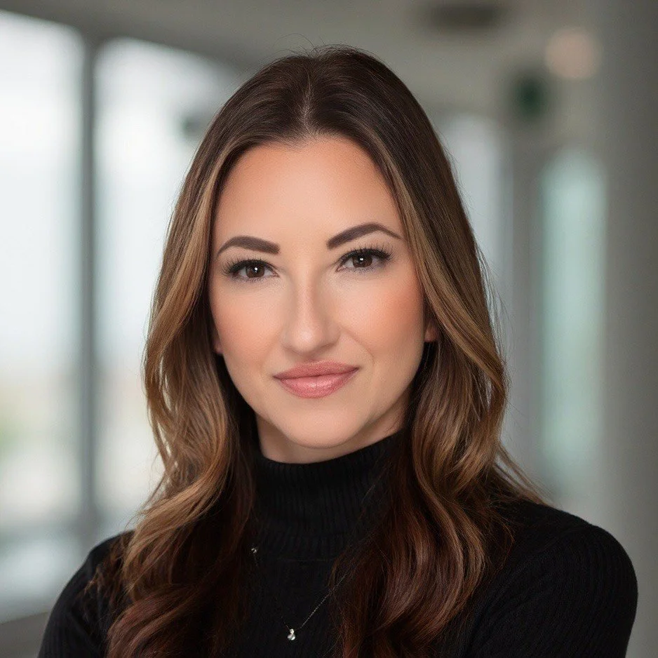 A woman with long wavy brown hair wearing a black turtleneck and a delicate necklace, smiling in an indoor setting with blurred windows in the background.