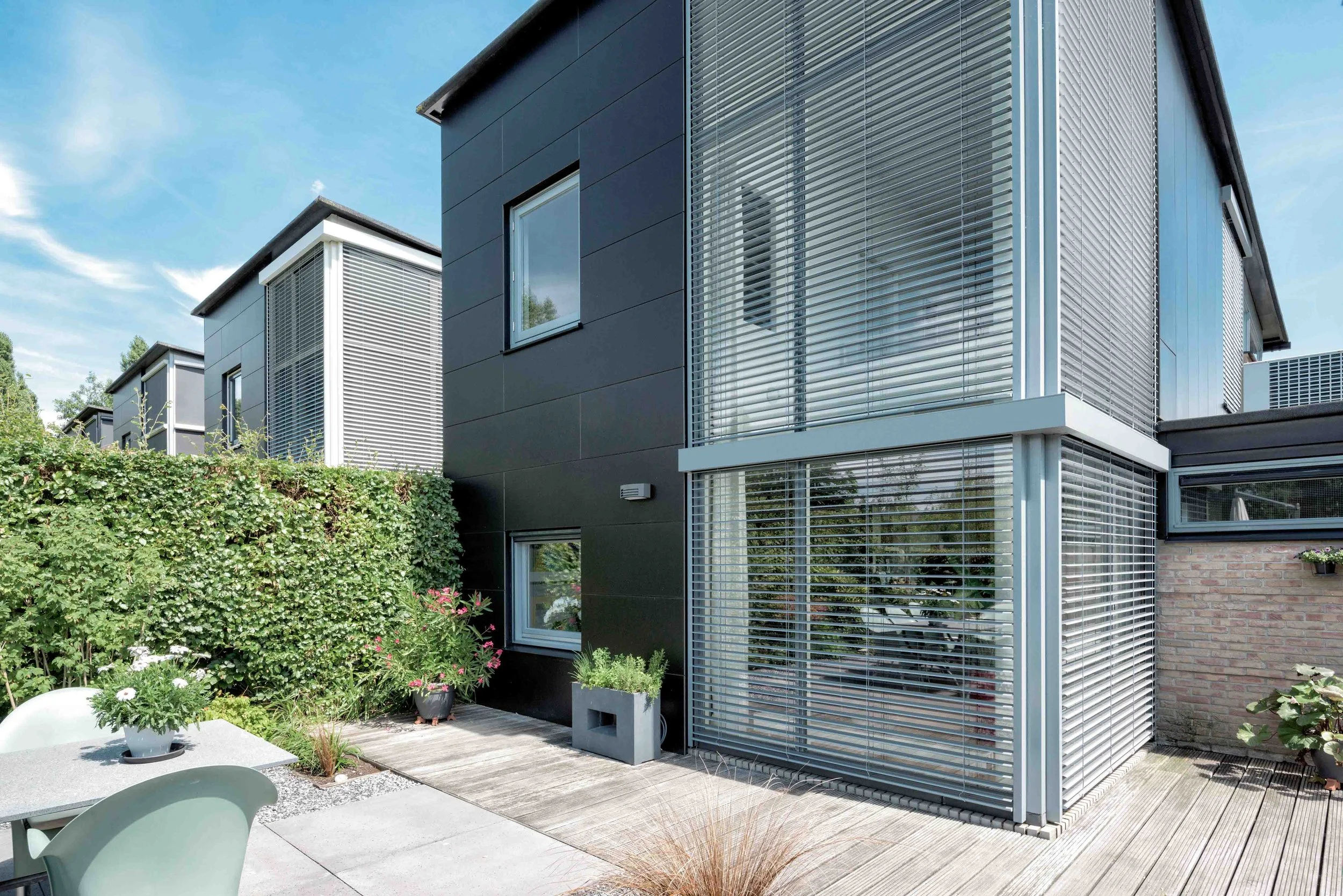 Moderne Wohnhäuser mit schwarzer und weißer Fassade, große Fenster mit Rollläden, gepflegte Garten- und Terrassenbereich bei sonnigem Wetter.