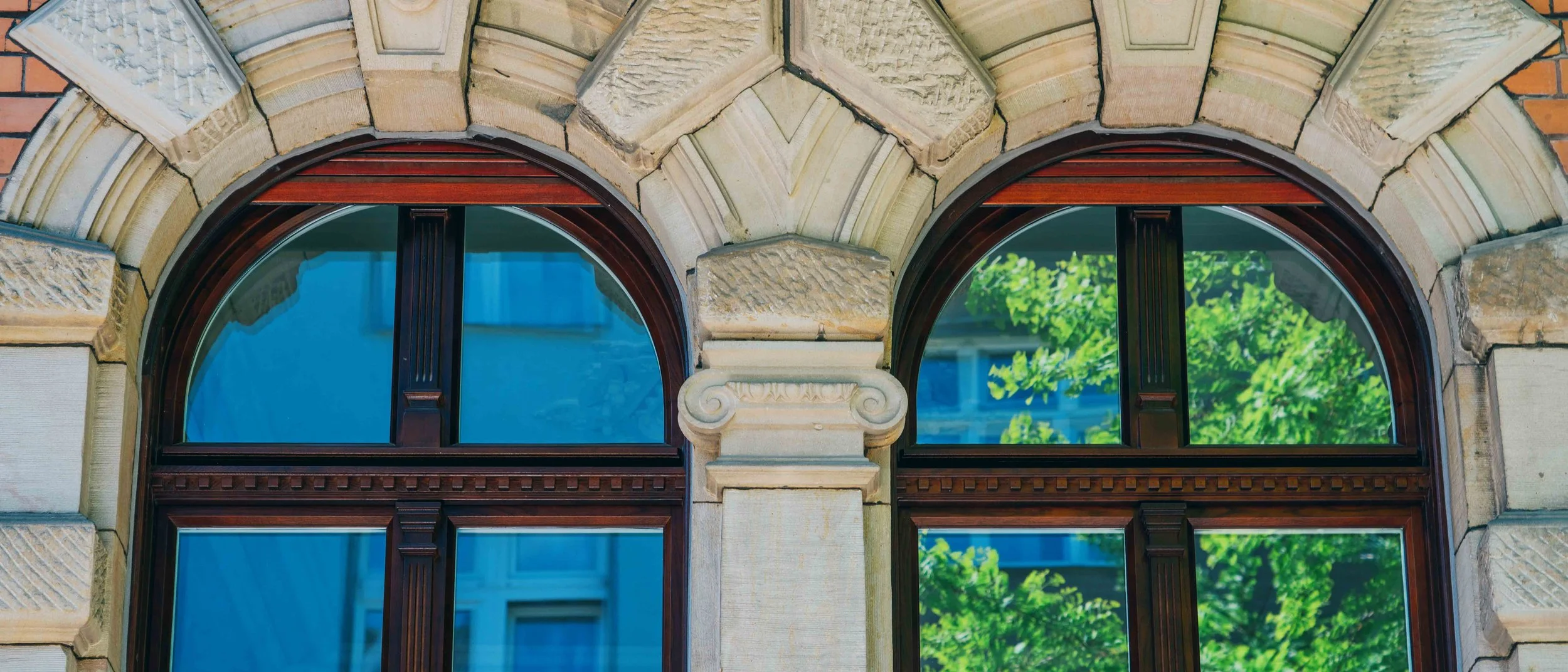 Zwei arched Fenster mit dunklem Holzrahmen, umgeben von historischer Steinarchitektur, mit reflektierendem Glas, das grüne Bäume draußen zeigt.