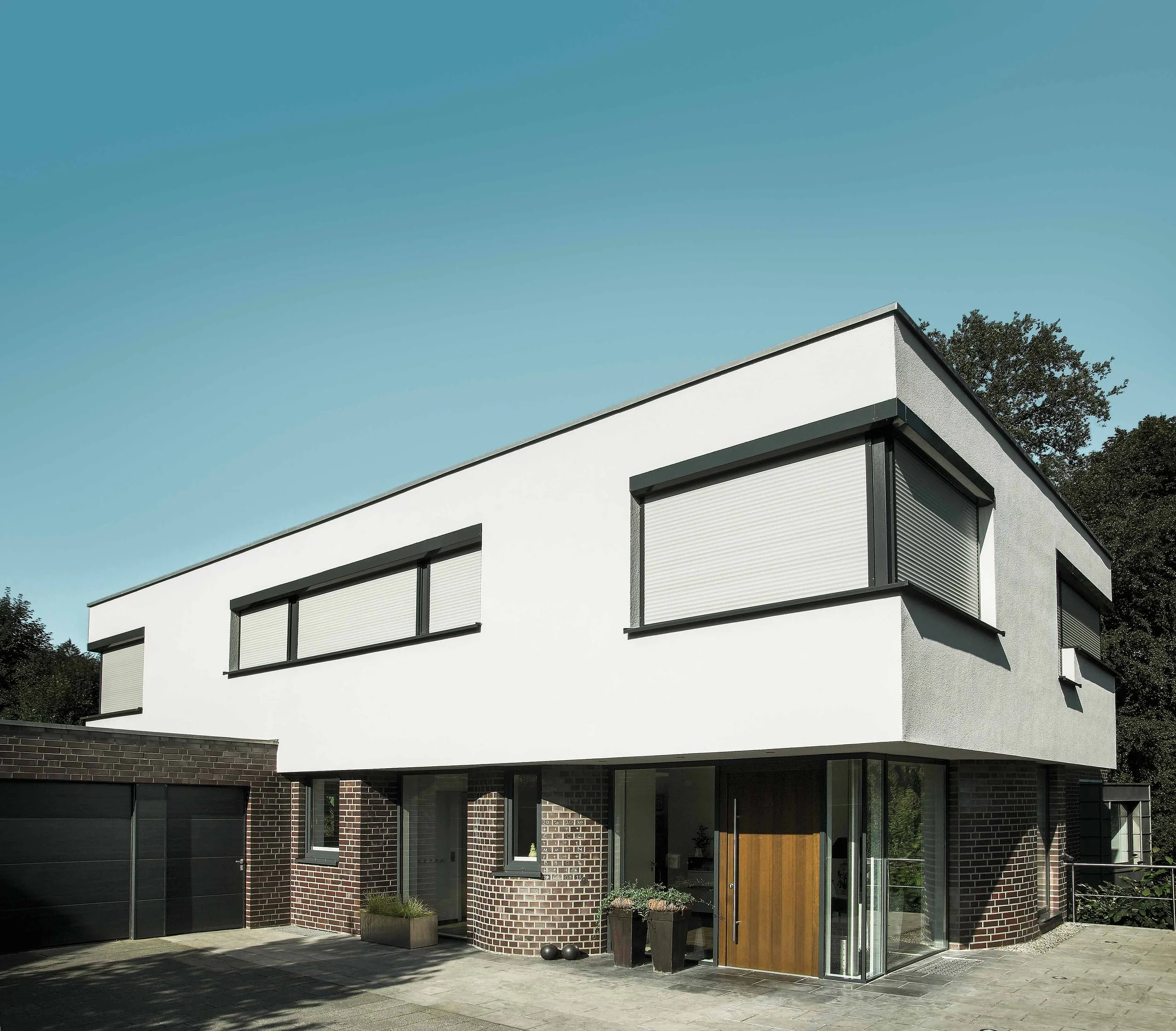 Modernes, mehrstöckiges Haus mit weißen und dunklen Elementen, große Fenster mit Rollläden, Eingang mit Holztreppe, Garage auf der linken Seite, umgeben von Bäumen, bei sonnigem Wetter.