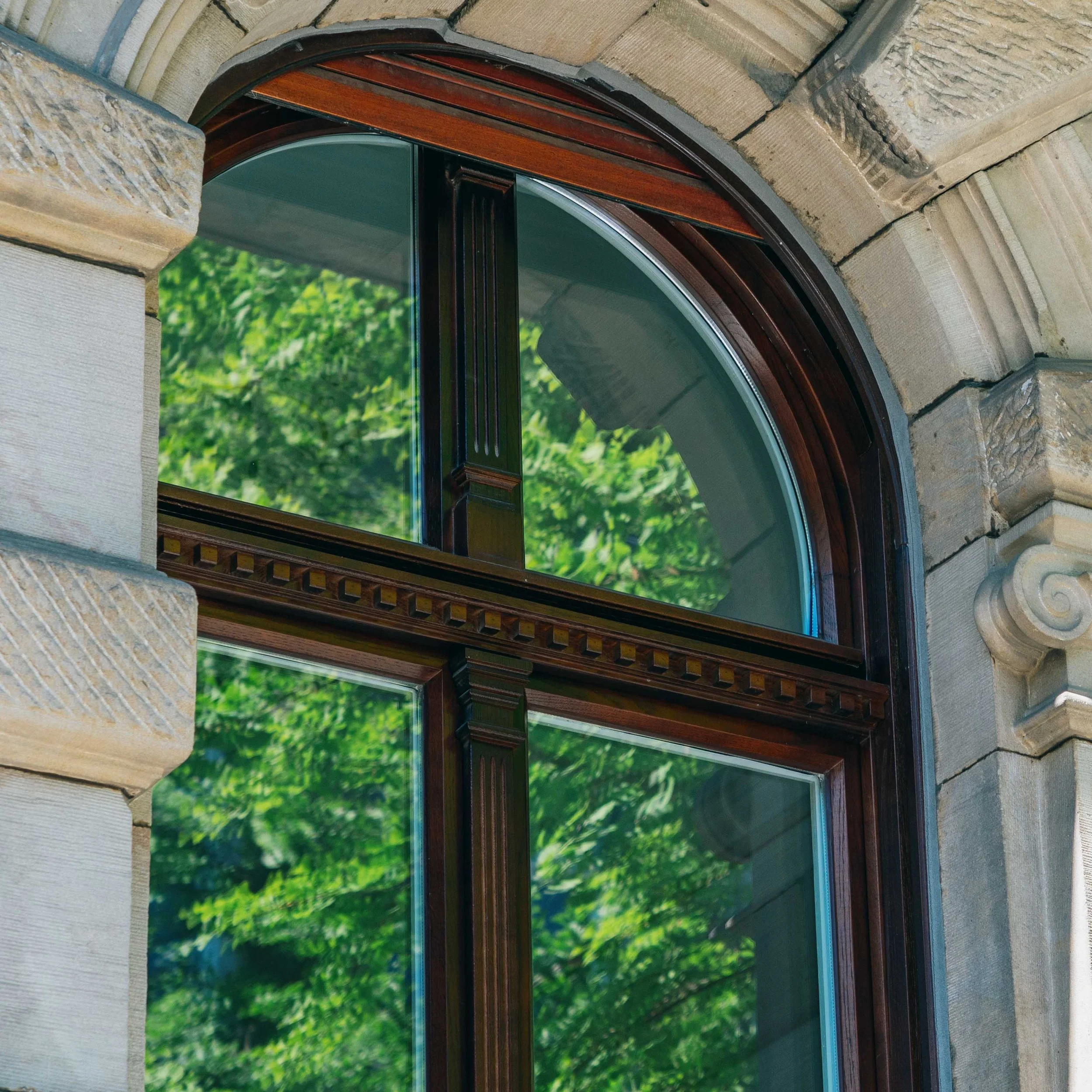 Ein bogenförmiges Fenster mit dunklem Holzrahmen vor einer Steinmauer, durch das grüne Blätter eines Baumes sichtbar sind.
