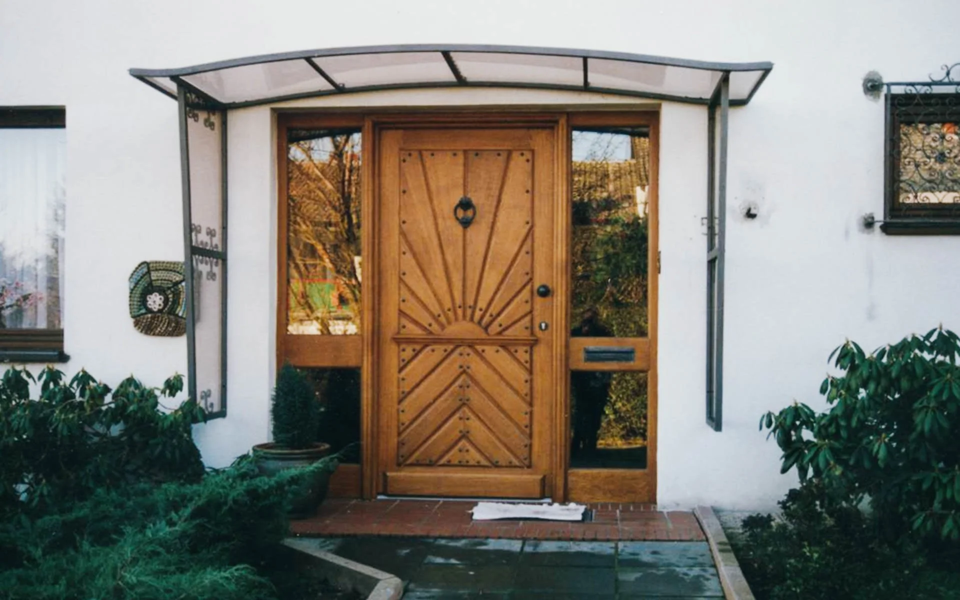 Holztür mit geometrischem Muster, umgeben von Fenstern, vor einem weißen Haus, mit Pflanzen und einem kleinen Teich im Eingangsbereich.