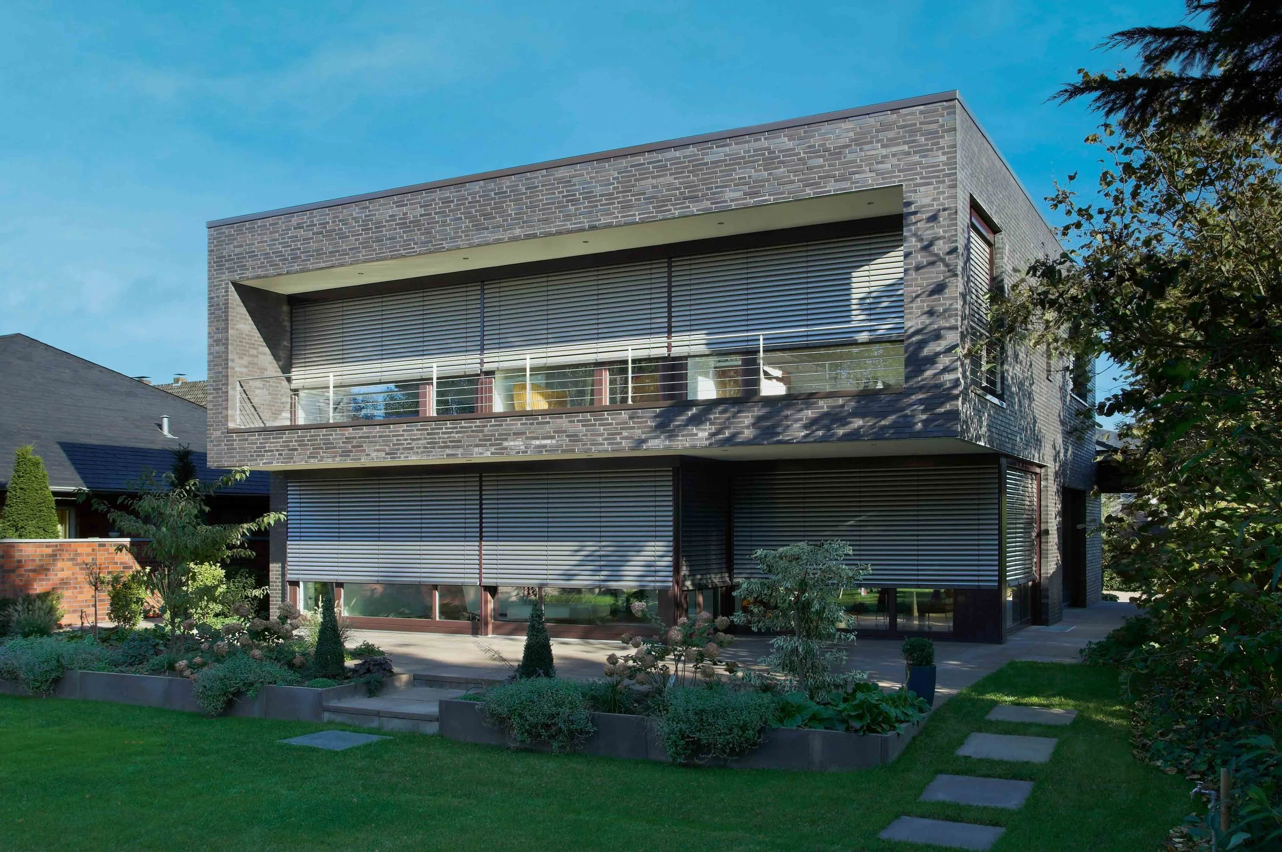Modernes zweistöckiges Haus mit dunklen Ziegelwänden und großen Fensterfronten, umgeben von einem gepflegten Garten mit Steinen und Büschen, bei sonnigem Himmel.