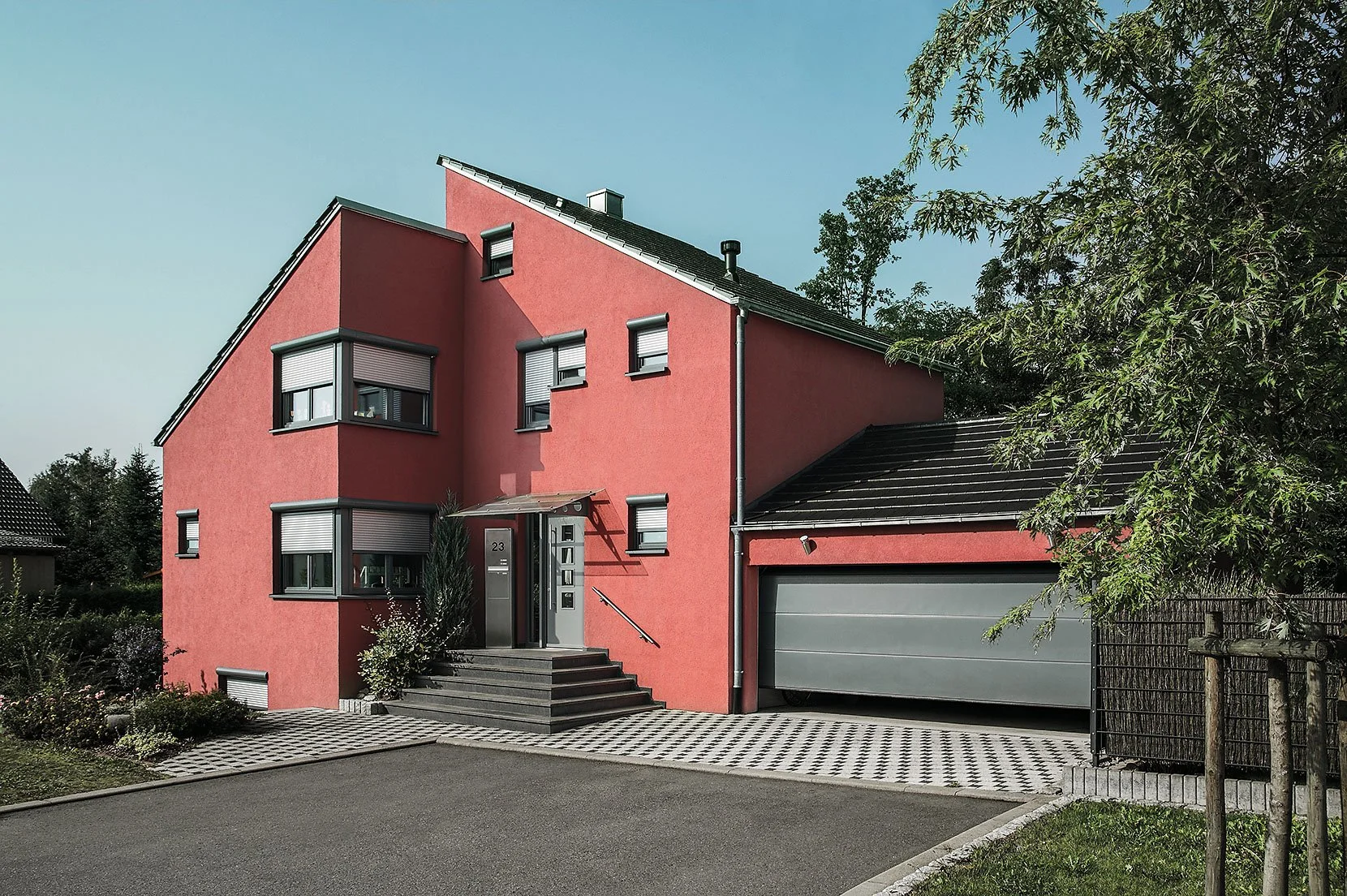 Modernes rotes Haus mit schwarzen Fenstern, Eingangstreppe und Garage, umgeben von Grünflächen.