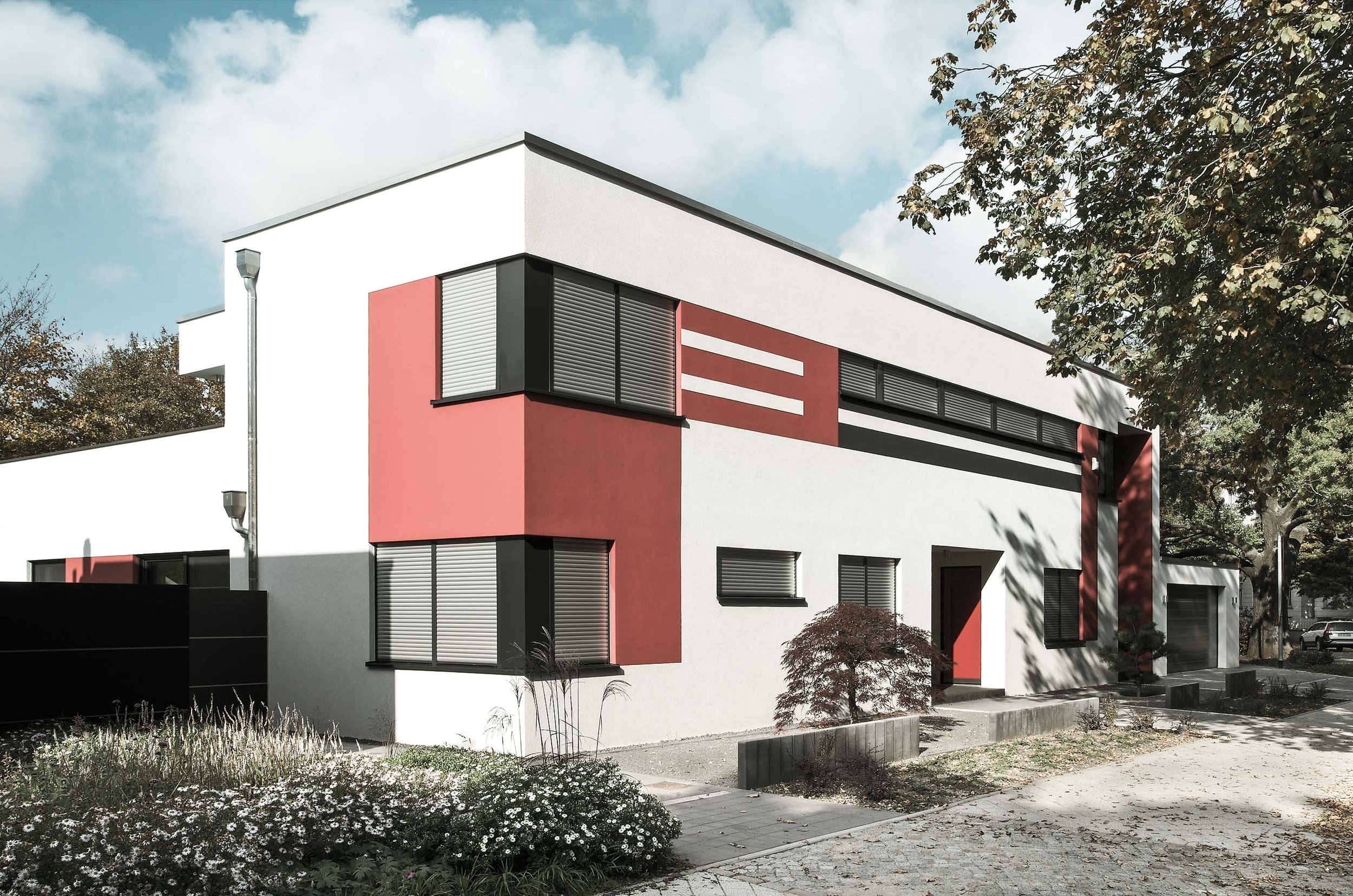Modernes, zweistöckiges Wohnhaus mit weißen und roten Fassaden, mehrere Fenster mit Rollläden, vor dem Eingang kleine Bäume und Blumen, umgeben von Bäumen, bei bewölktem Himmel.