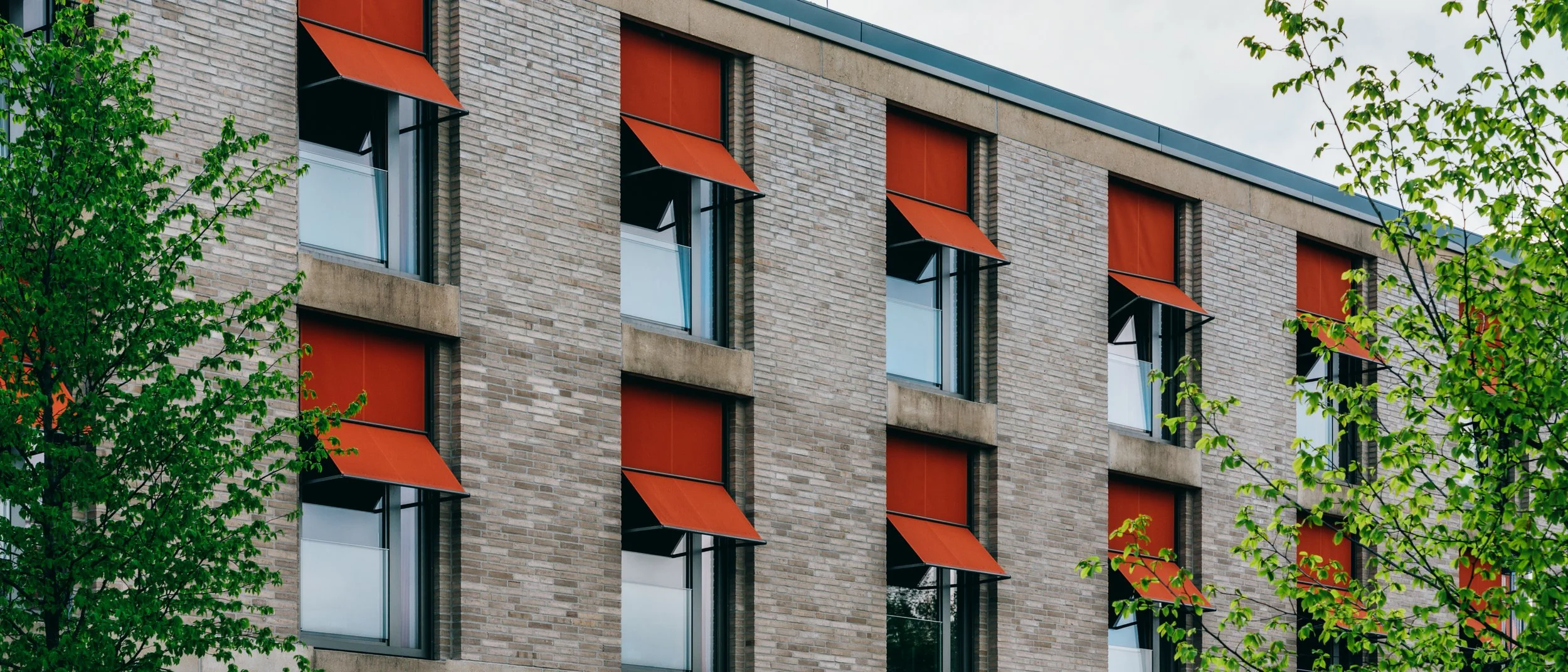 Mehrstöckiges modernes Gebäude mit roten Awnings über den Fenstern, umgeben von grünen Bäumen.