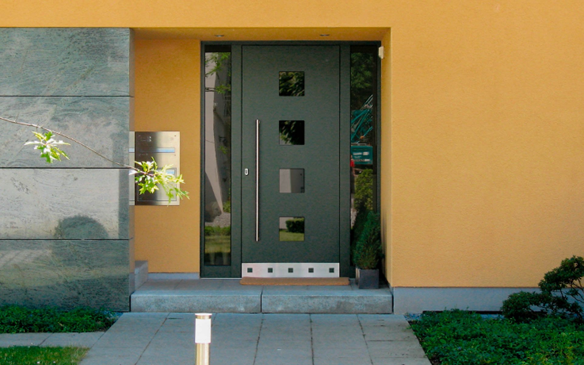 Moderner Hauseingang mit dunkler Haustür, Fenster und gelber Wand im Hintergrund, flankiert von grünen Pflanzen und einer Gehweg-Terrasse.