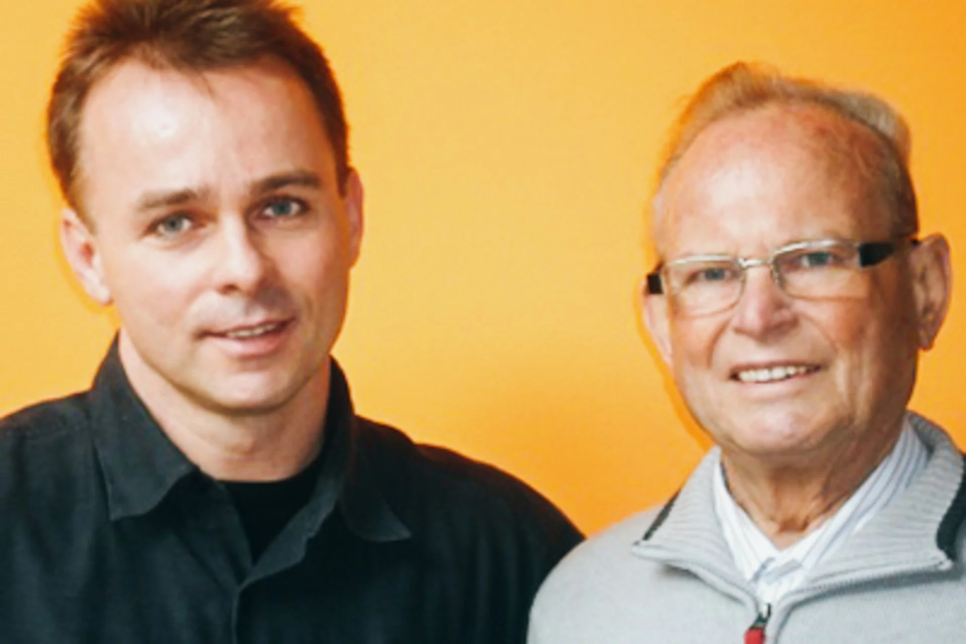Zwei Männer vor einem orangefarbenen Hintergrund, lächelnd in die Kamera schaun.
