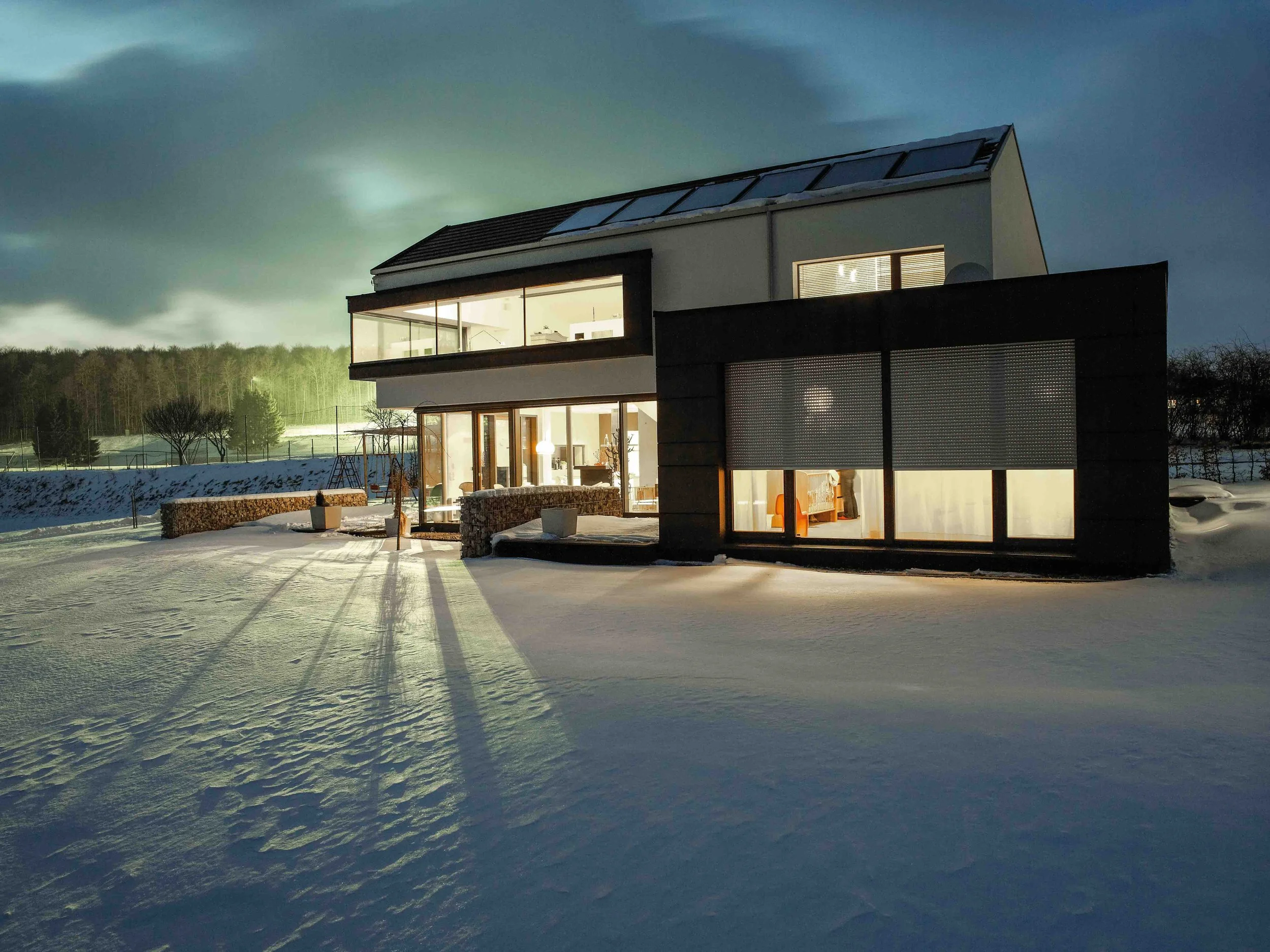 Moderne, zweistöckige Villa mit großen Fenstern und Solaranlagen auf dem Dach, umgeben von Schnee bei Nacht, beleuchtet im Innenraum.
